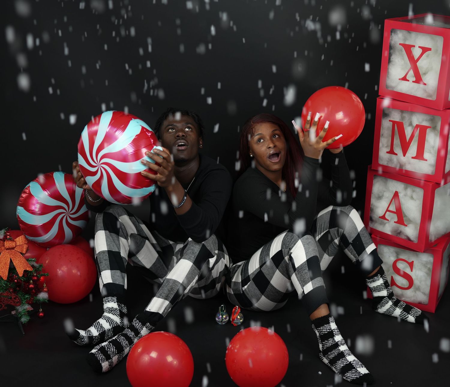 Couple in matching plaid pajamas hold balloons beside XMAS blocks with simulated snow effect.