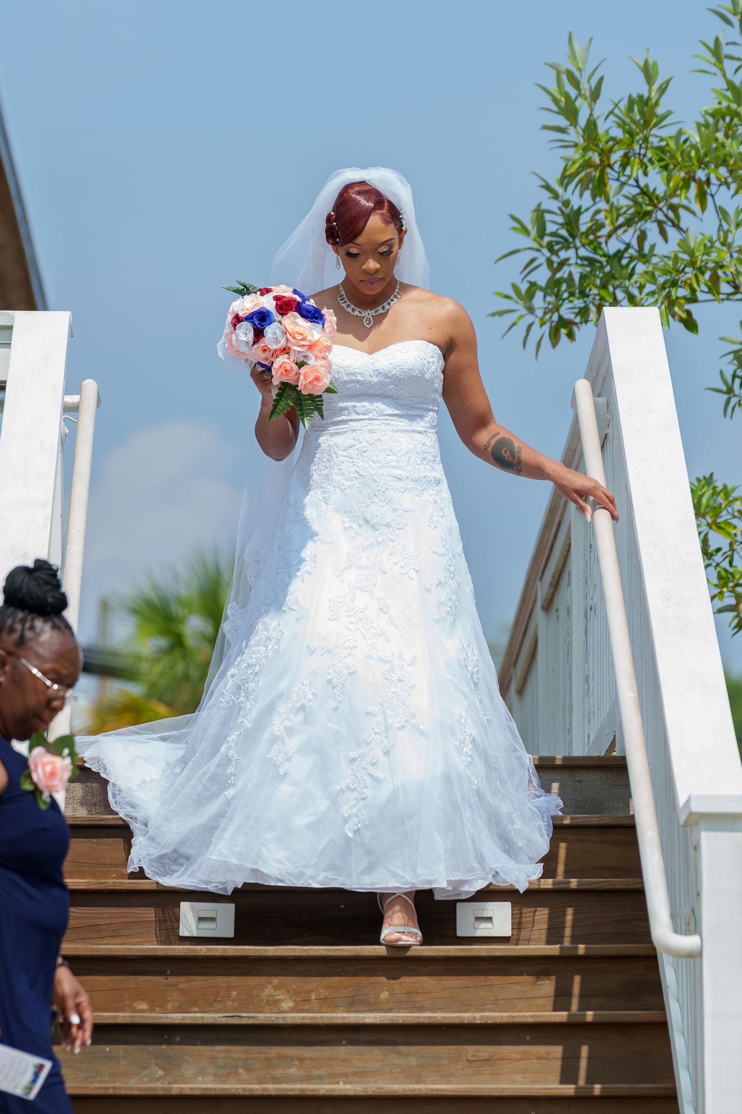 Elegant bride in strapless white gown holding colorful bouquet descending outdoor wooden stairs.
