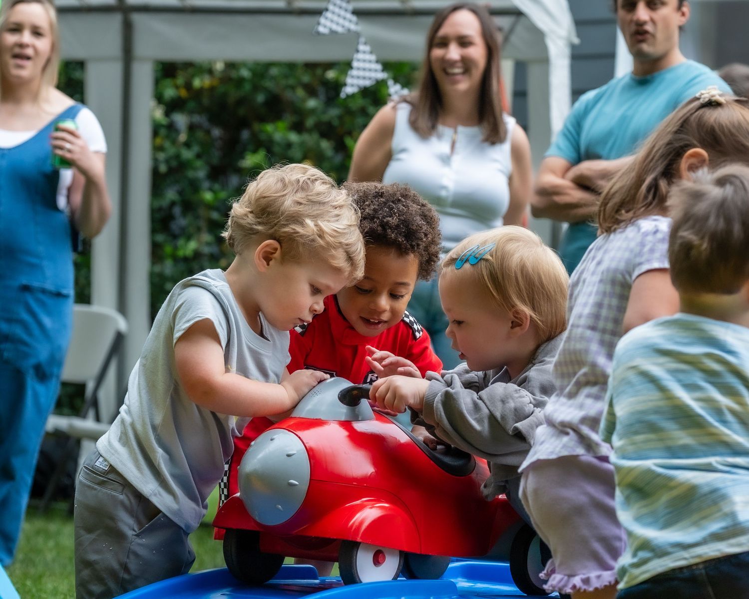 Group of young children gathered around a red toy car at an outdoor birthday party.