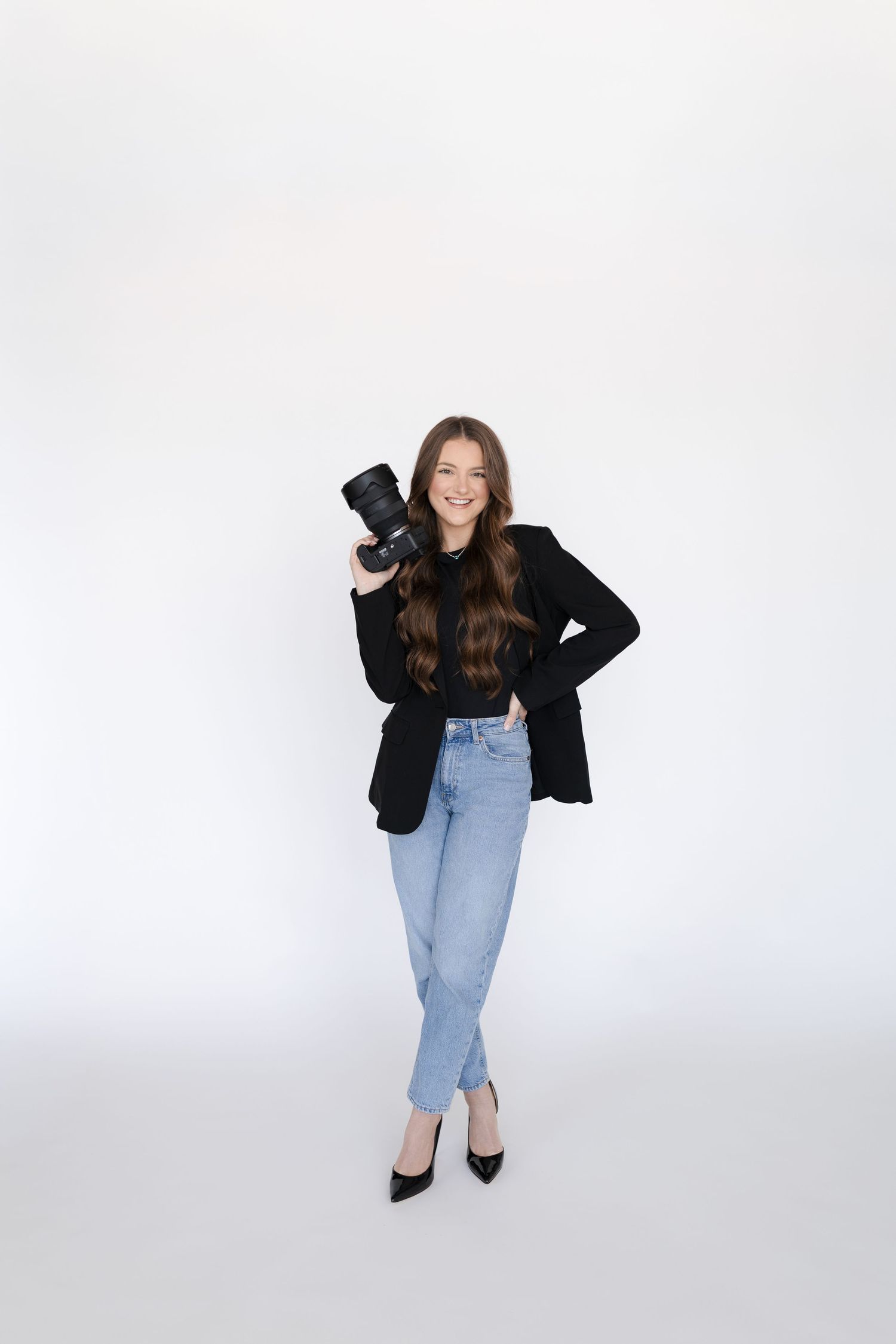 Smiling female photographer in black blazer and jeans holding a camera against a white background.