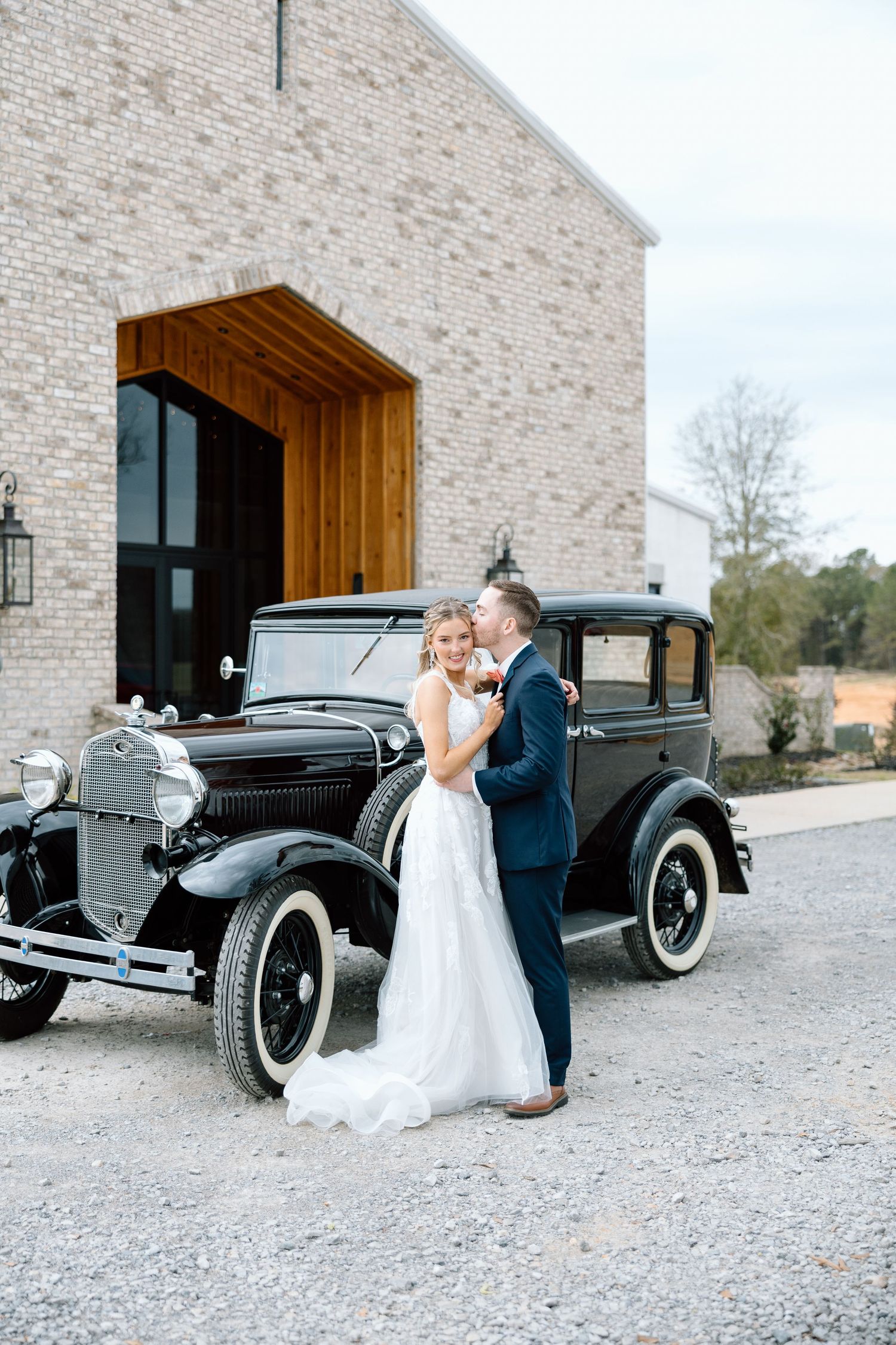 Bride and groom posing beside vintage black car outside rustic brick wedding venue.