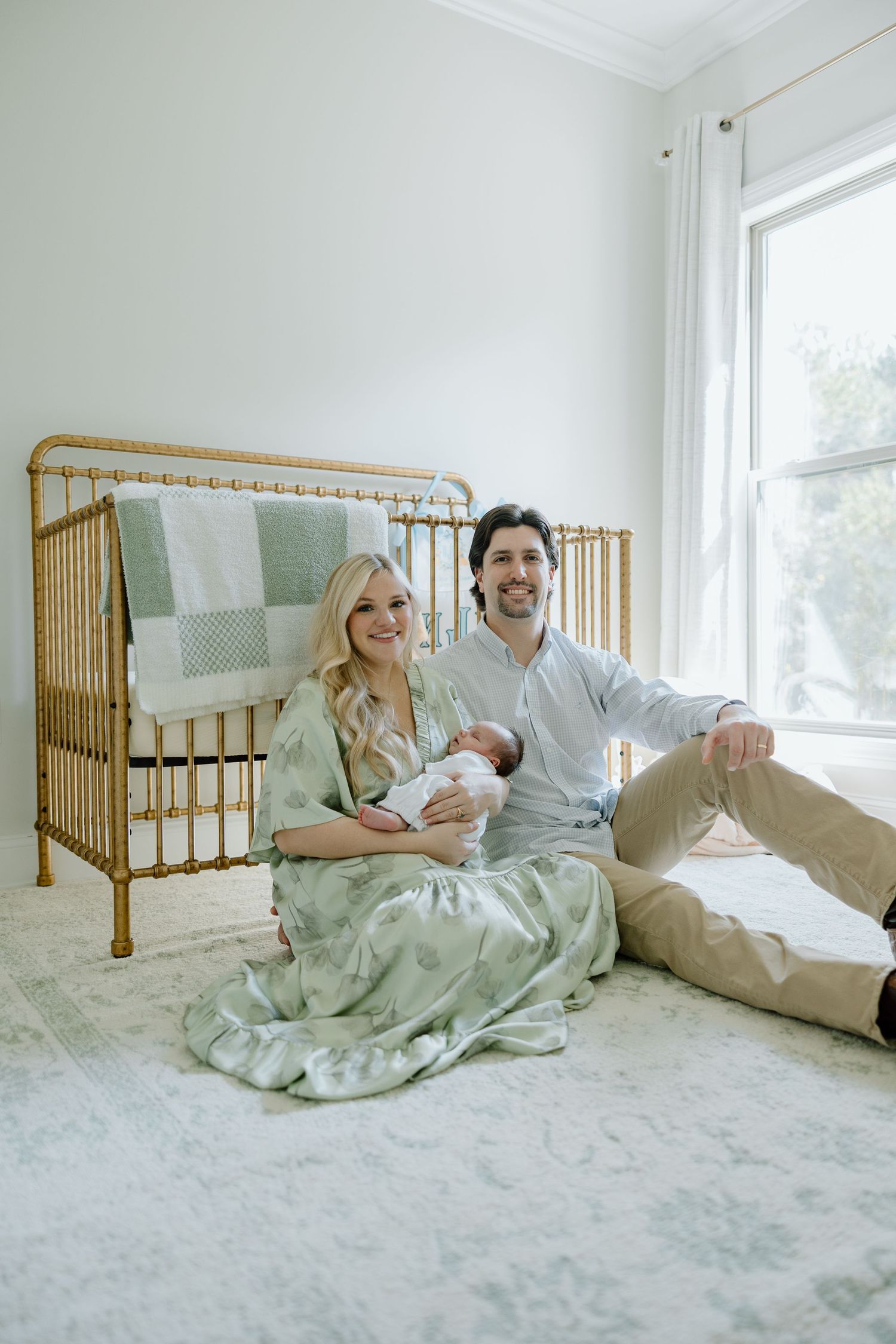 New parents sitting on nursery floor with newborn baby beside a gold vintage crib.