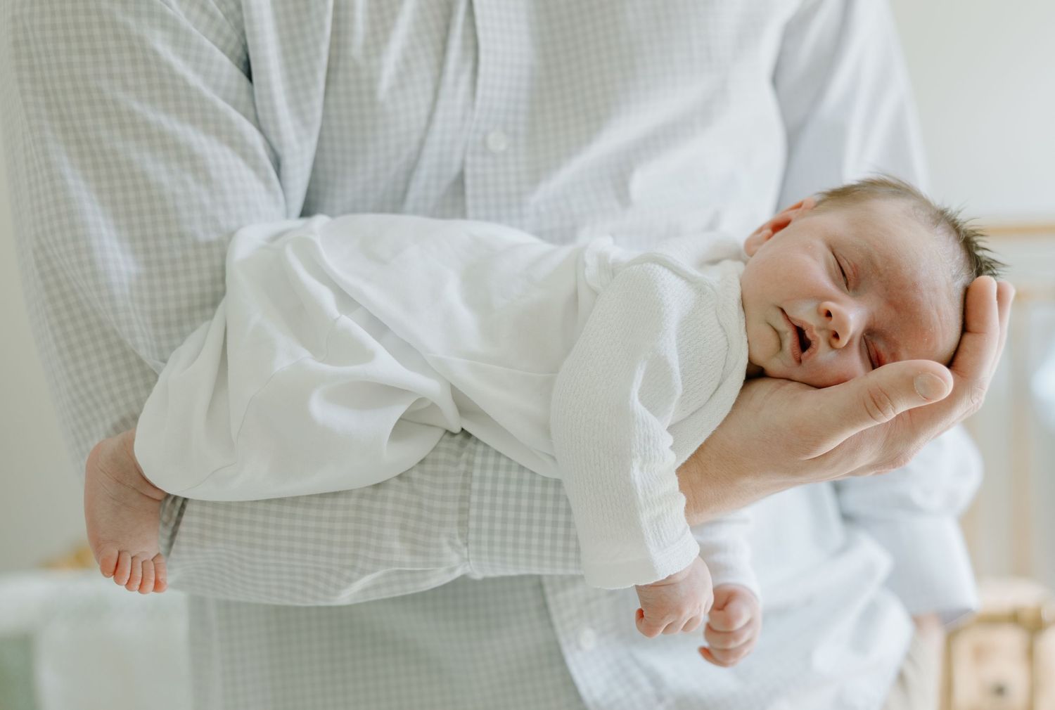 Father in blue shirt gently cradling sleeping swaddled newborn baby against his chest.