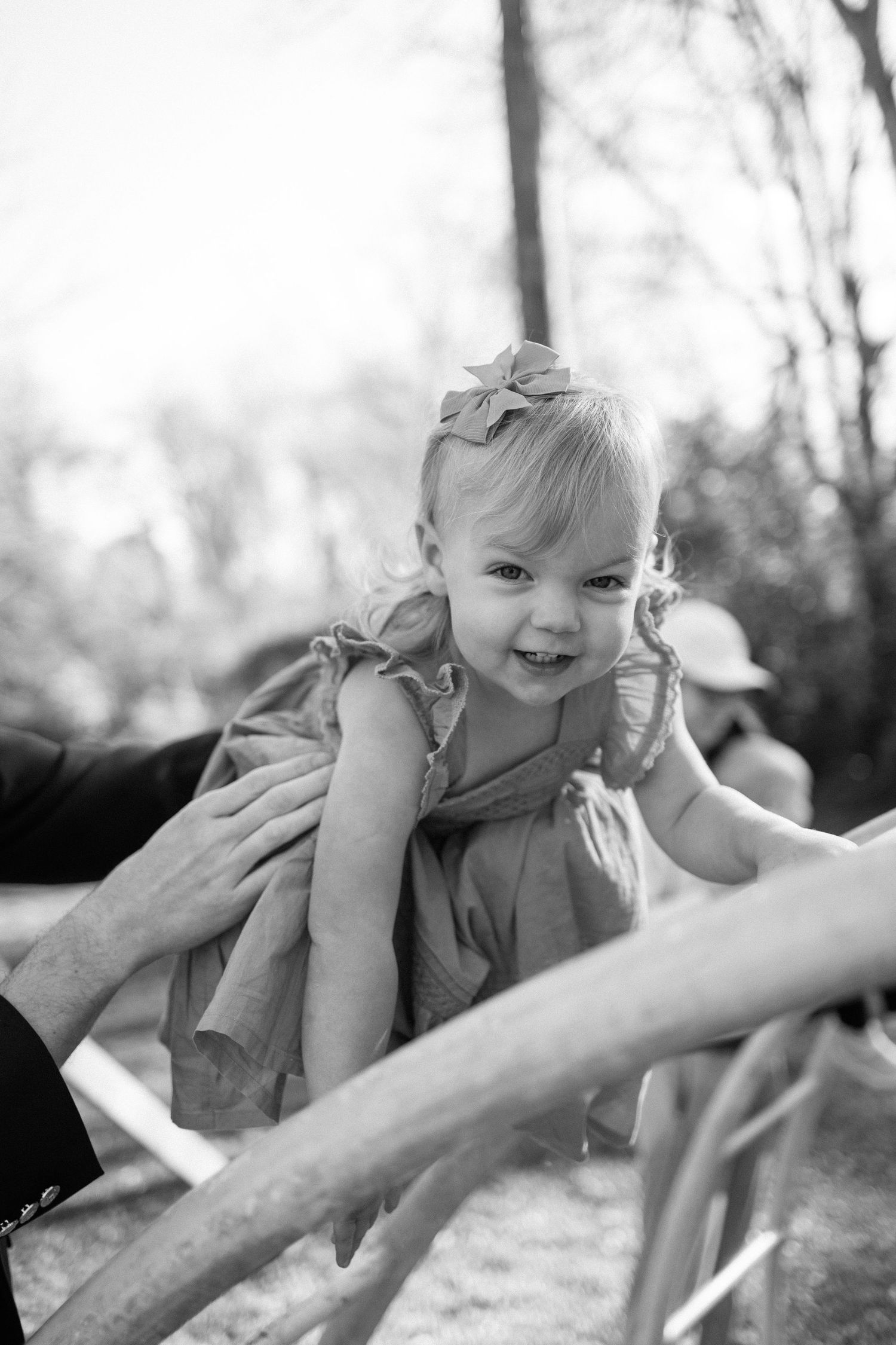 Smiling toddler girl with flower headband being lifted by adult hands at outdoor playground.