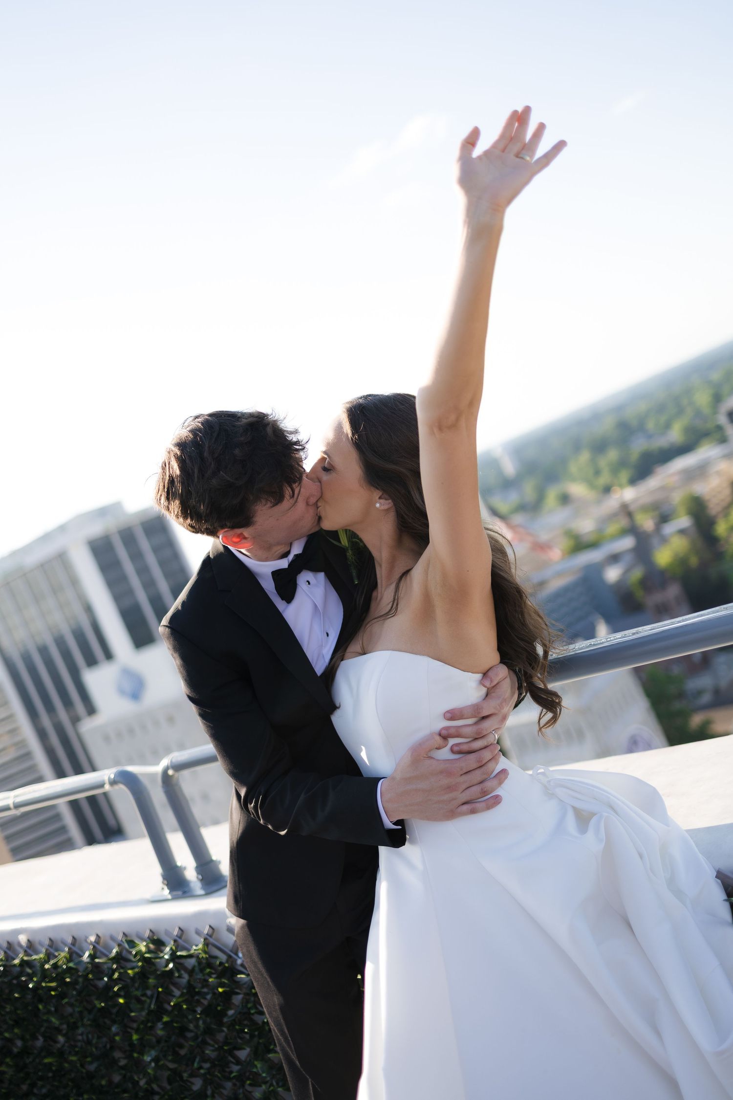 Bride and groom share a romantic kiss on a rooftop, bride raising her hand joyfully in a white gown and tuxedo.