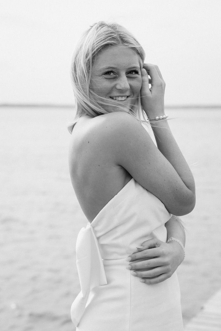 Smiling blonde woman in white halter dress standing near water in black and white.