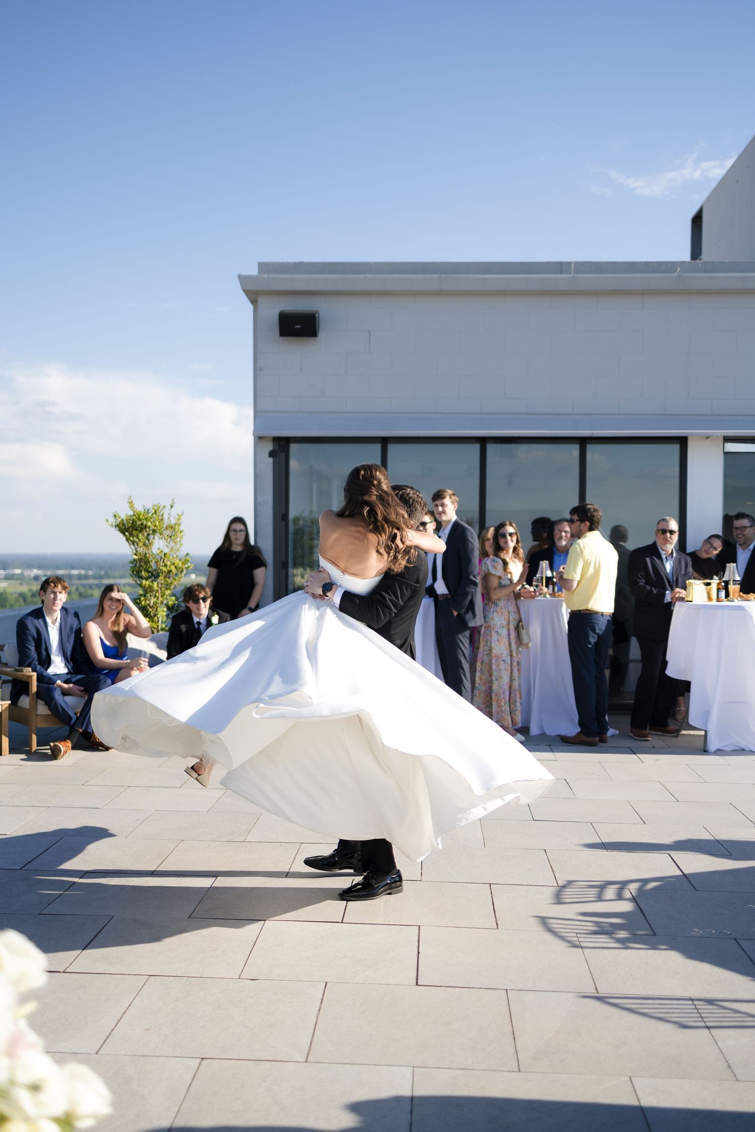 Bride and groom share first dance on rooftop venue terrace with guests watching under clear blue sky.