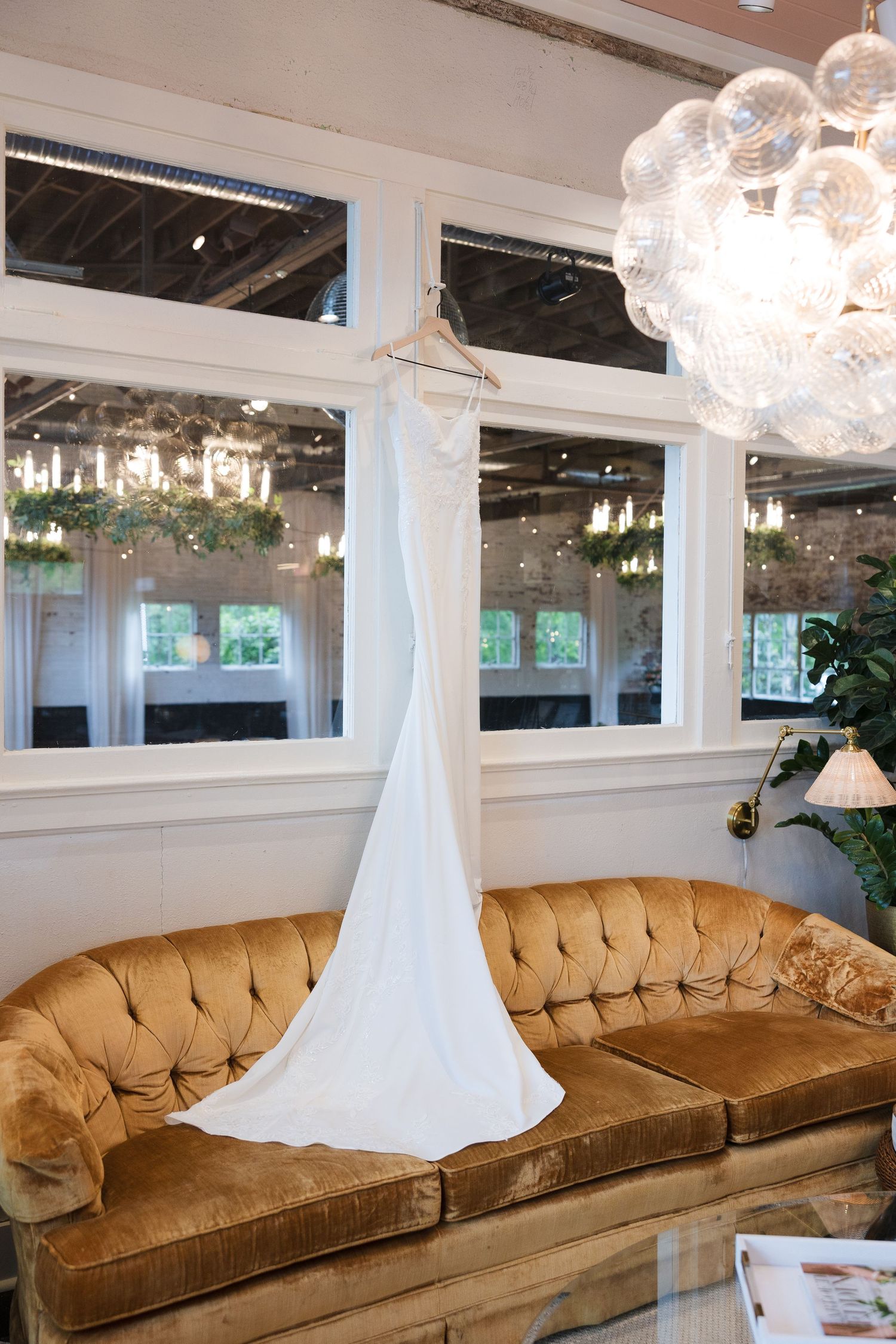 White wedding dress hanging above a tufted gold velvet sofa in an elegant bridal boutique with chandelier lighting.