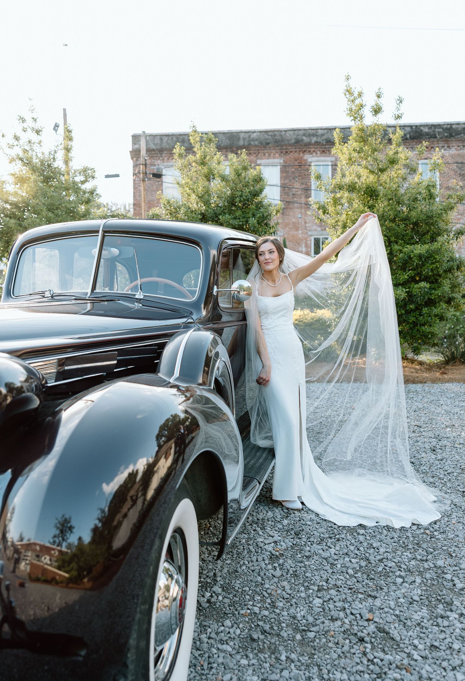 Bride in fitted white gown with cathedral veil poses beside vintage black classic car outdoors.