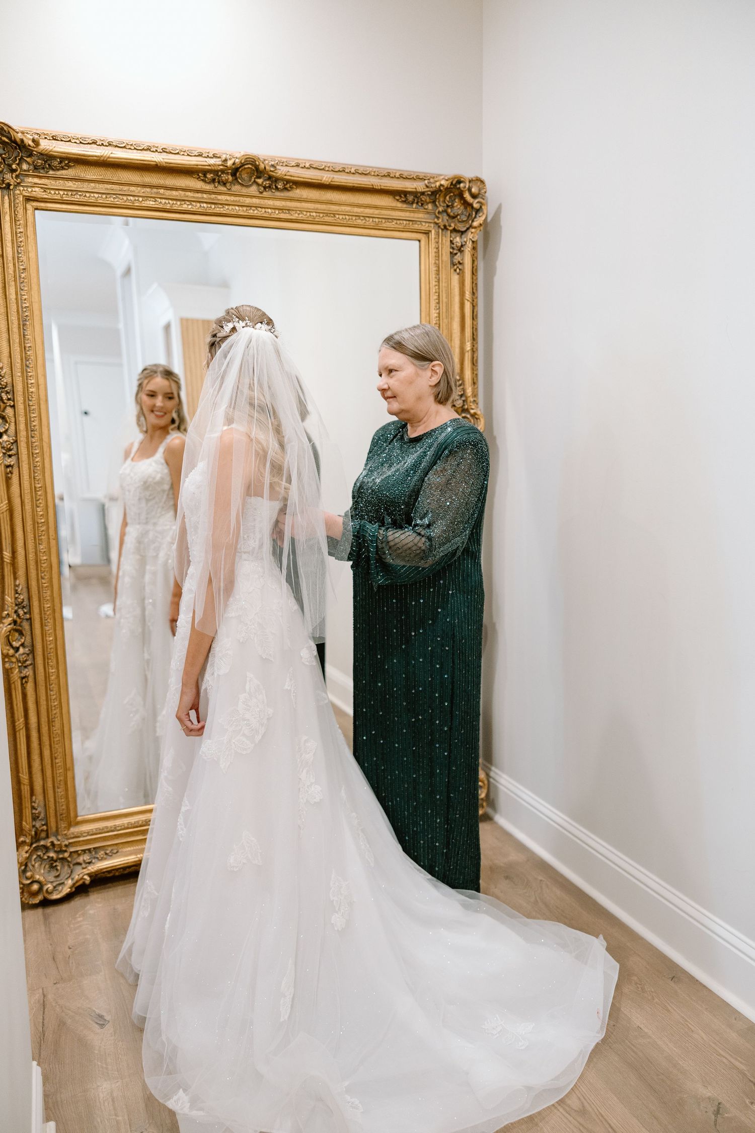 Bride in lace wedding gown with veil standing before gold mirror with mother.
