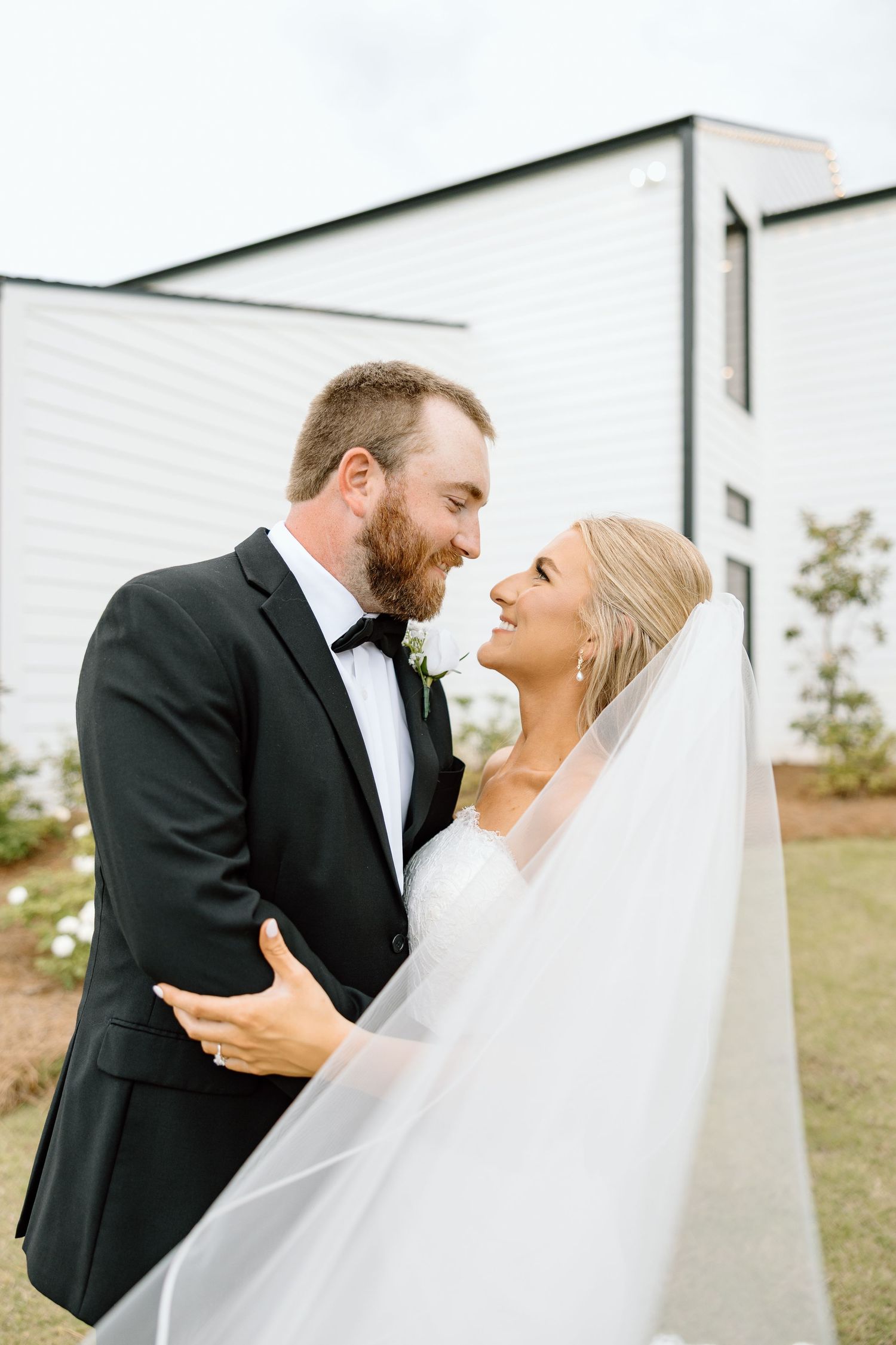 Bride and groom share an intimate moment with flowing veil outside white modern wedding venue.
