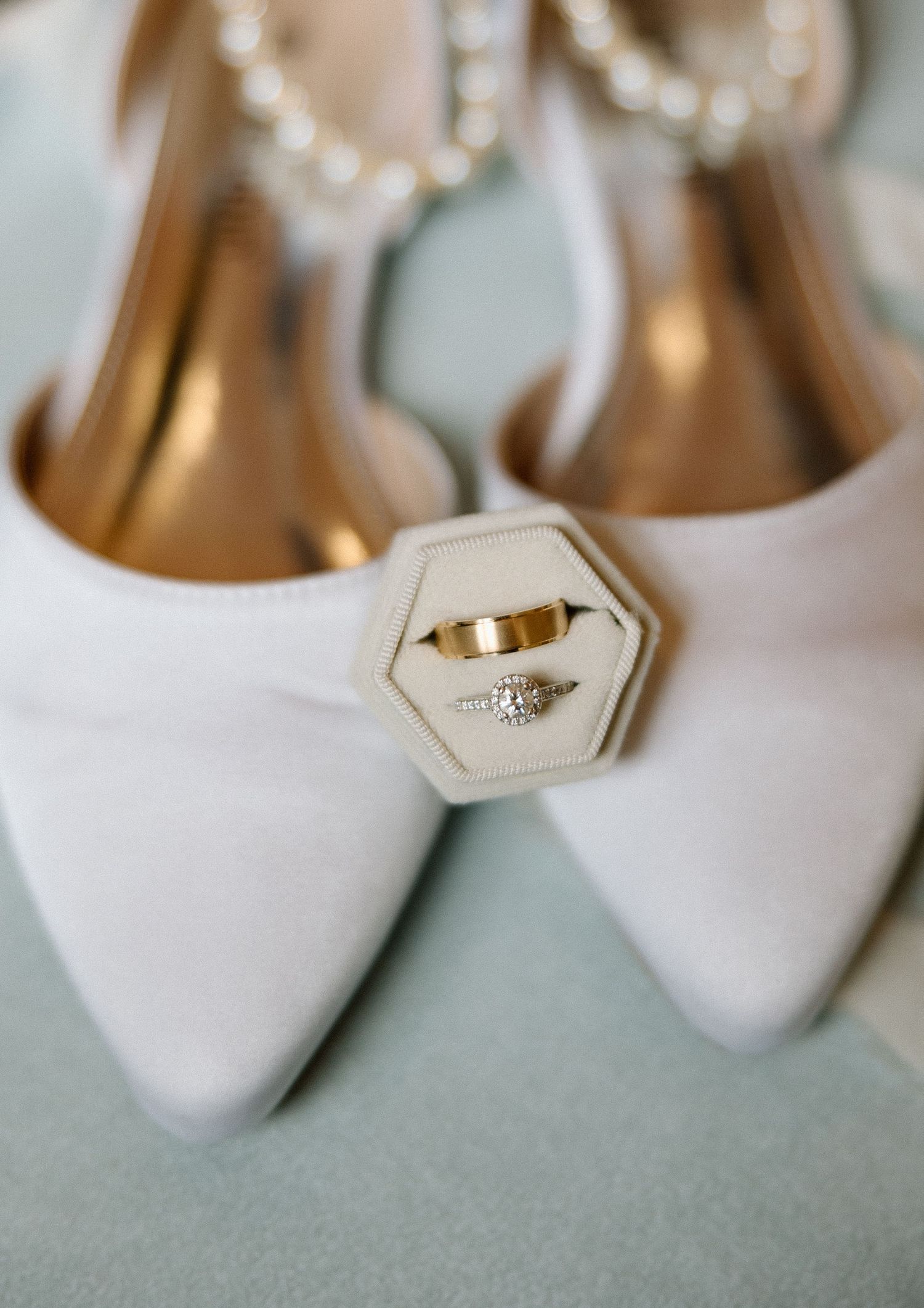 Gold wedding rings displayed in hexagon box on white bridal heels with pearl ankle straps.
