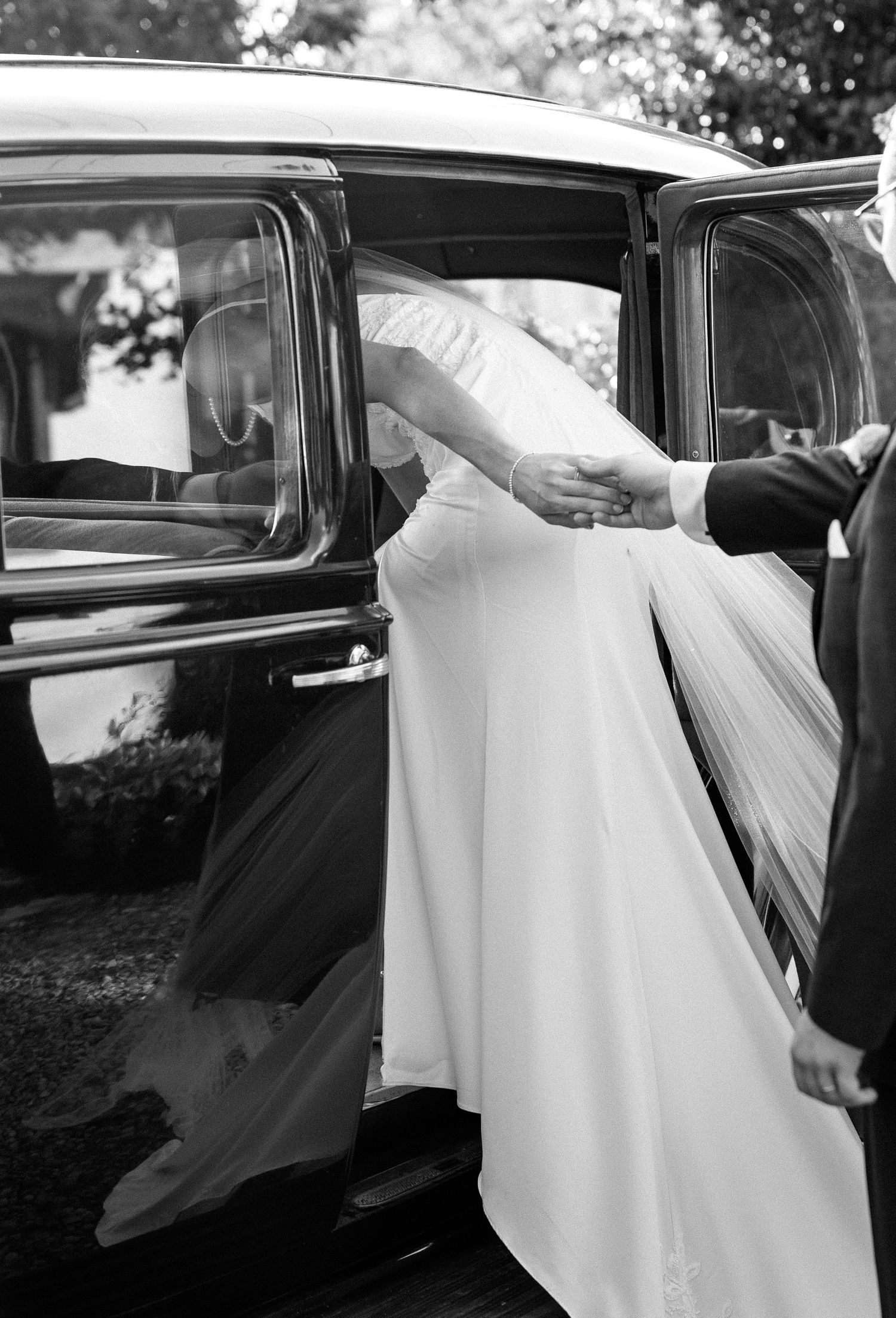 Bride in white gown being helped into a classic black car by groom, black and white wedding photo.