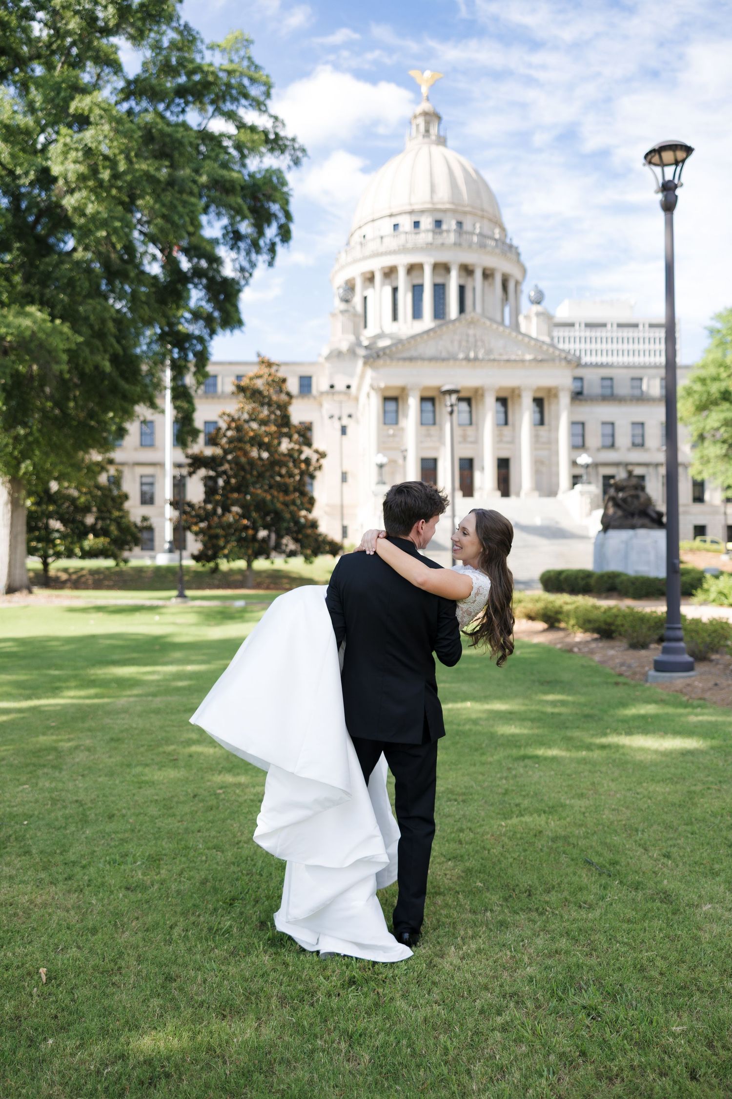 Groom lifting bride in white gown outside Mississippi State Capitol building on sunny wedding day.