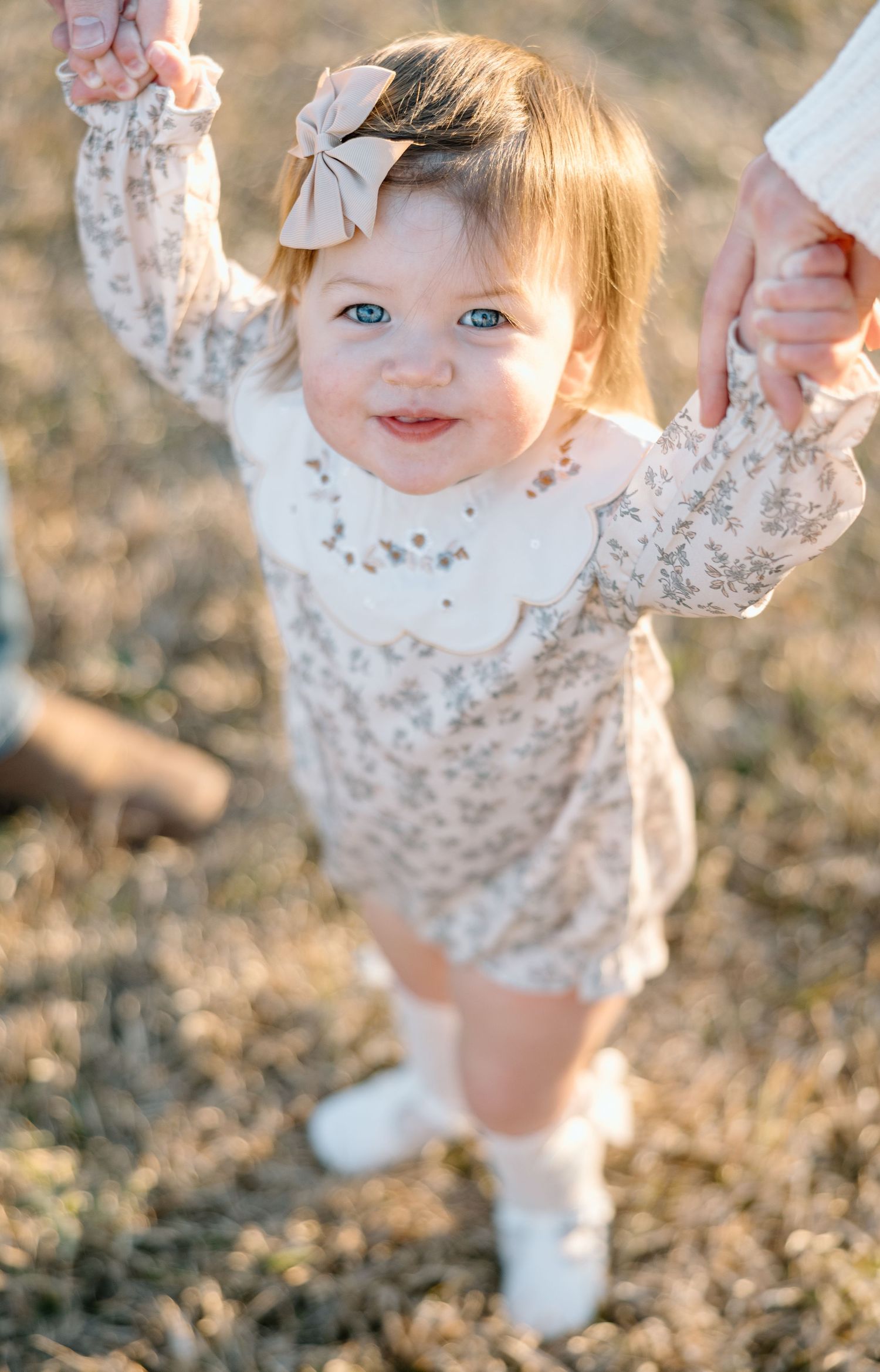 Adorable smiling baby girl in floral romper with bow headband being helped to walk outdoors.
