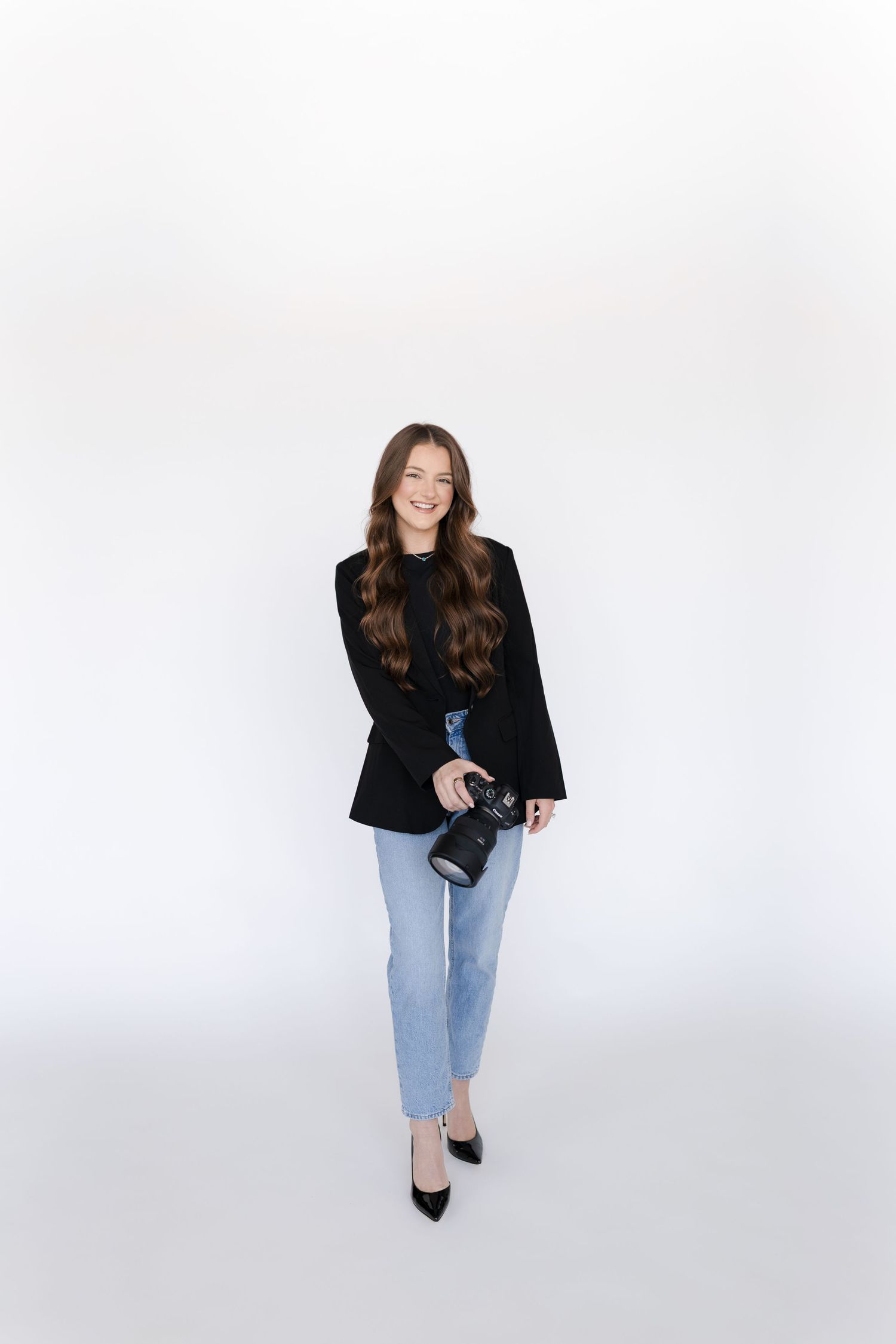 Smiling woman with long brown hair holding a camera, wearing a black blazer and light blue jeans on white background.