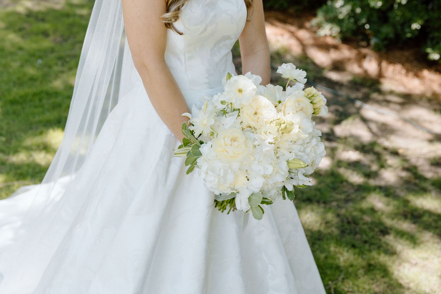 Bride in strapless white gown holding elegant white floral bouquet with veil outdoors.