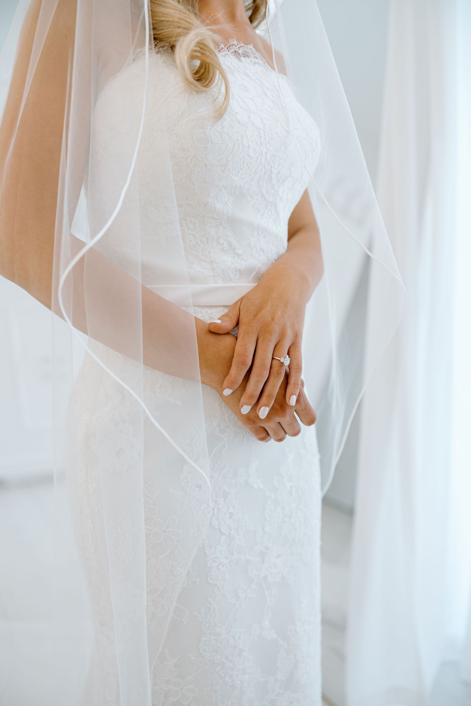 Bride in white lace gown and veil clasping hands showing diamond wedding ring.