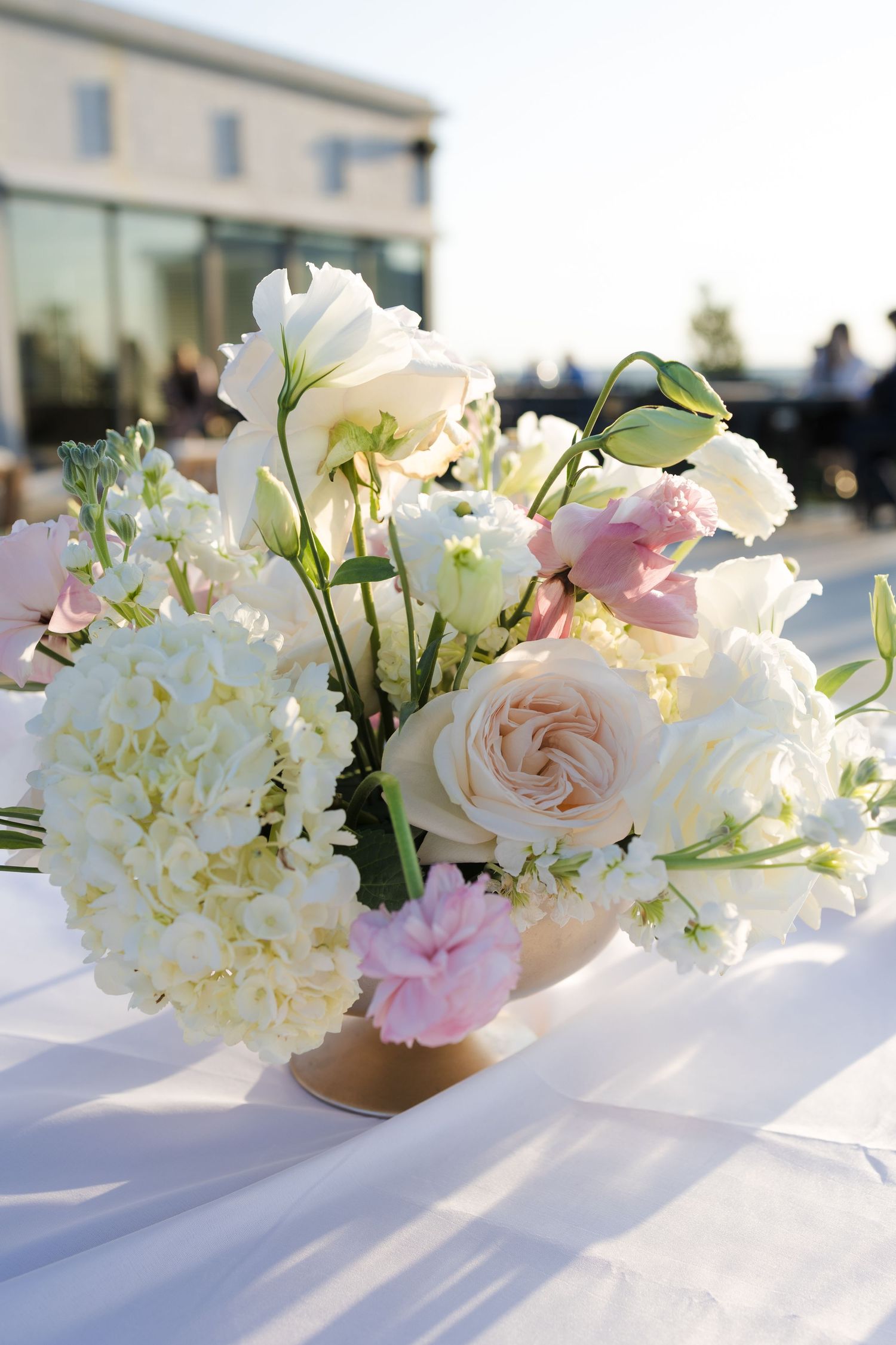 Elegant wedding centerpiece with white hydrangeas, blush roses, and pink lisianthus in a gold vase on white tablecloth.