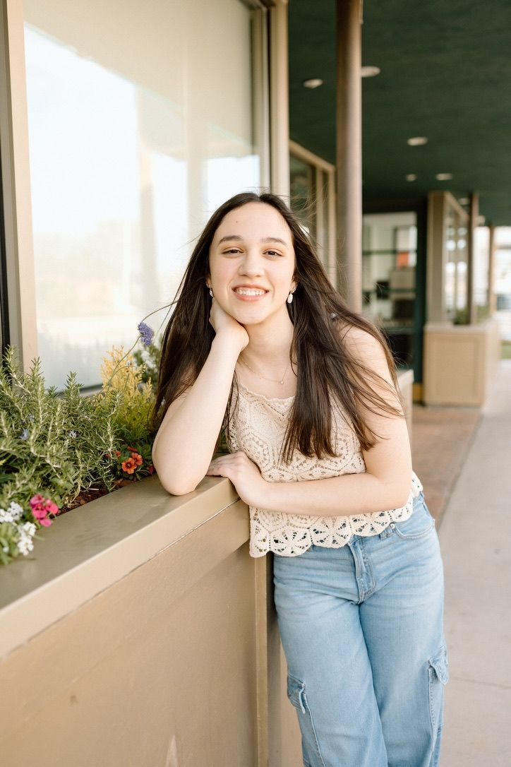 Smiling teenage girl with dark hair leaning on flower box outside storefront.