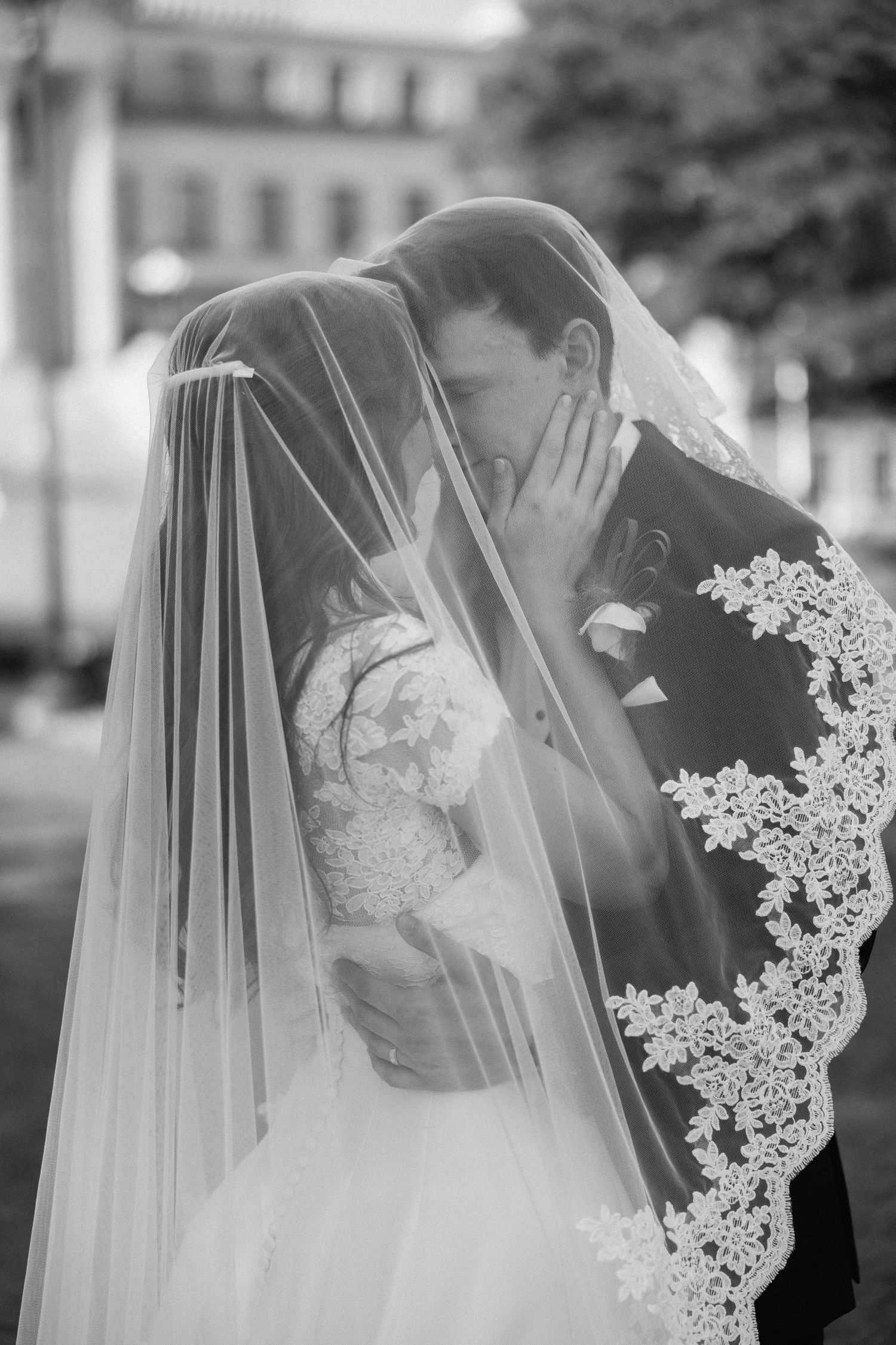 Black and white photo of bride and groom sharing a kiss under a lace wedding veil outdoors.