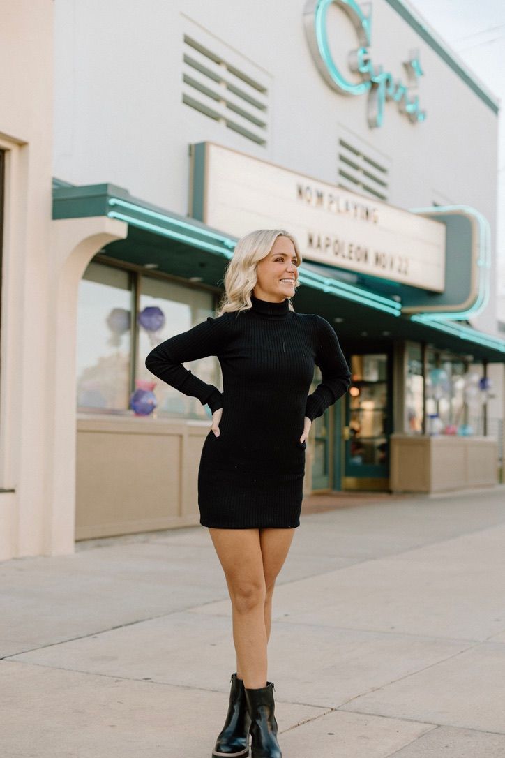 Blonde woman in black turtleneck mini dress posing outside vintage movie theater.