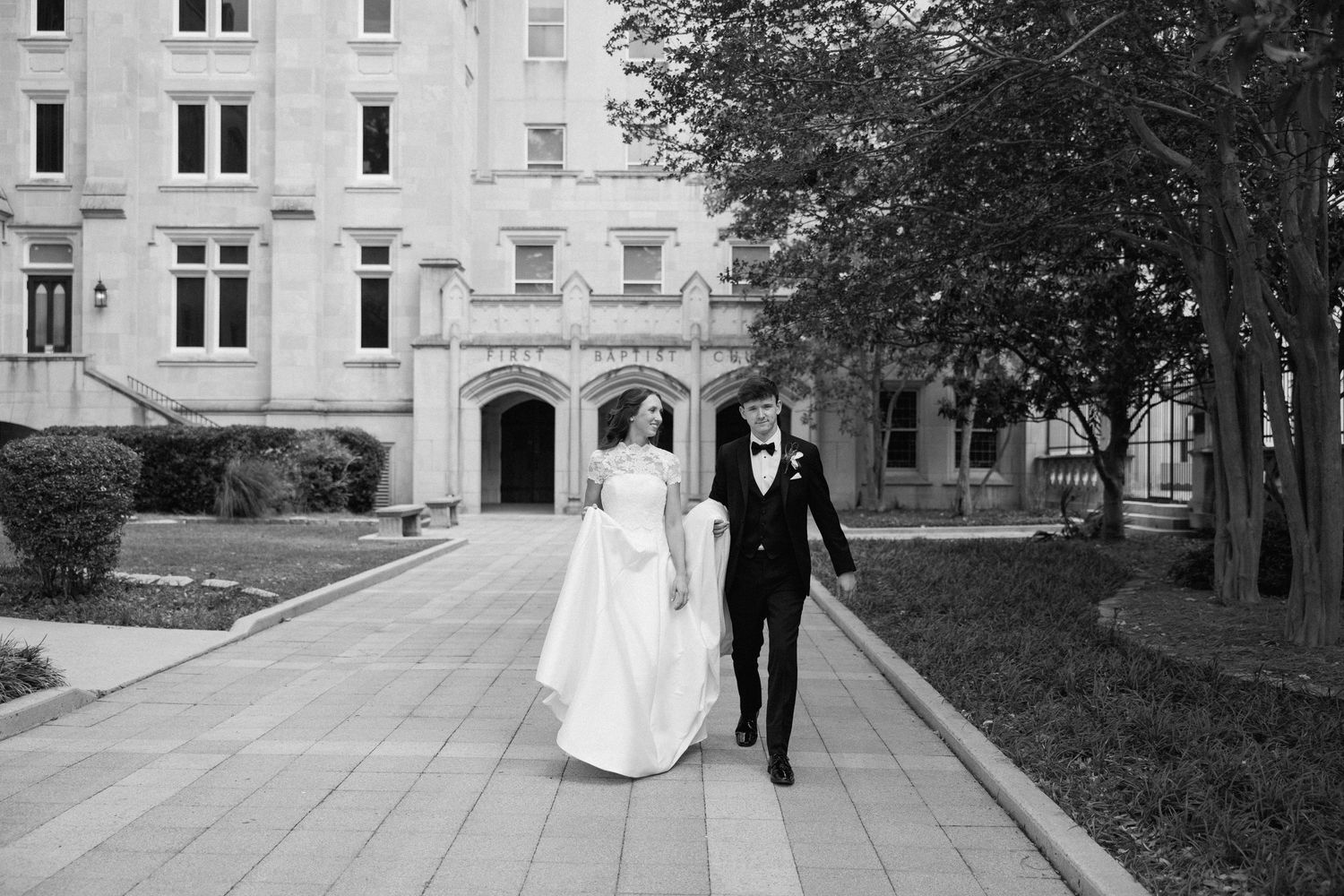 Black and white photo of a bride and groom walking hand in hand outside a grand historic building on their wedding day.