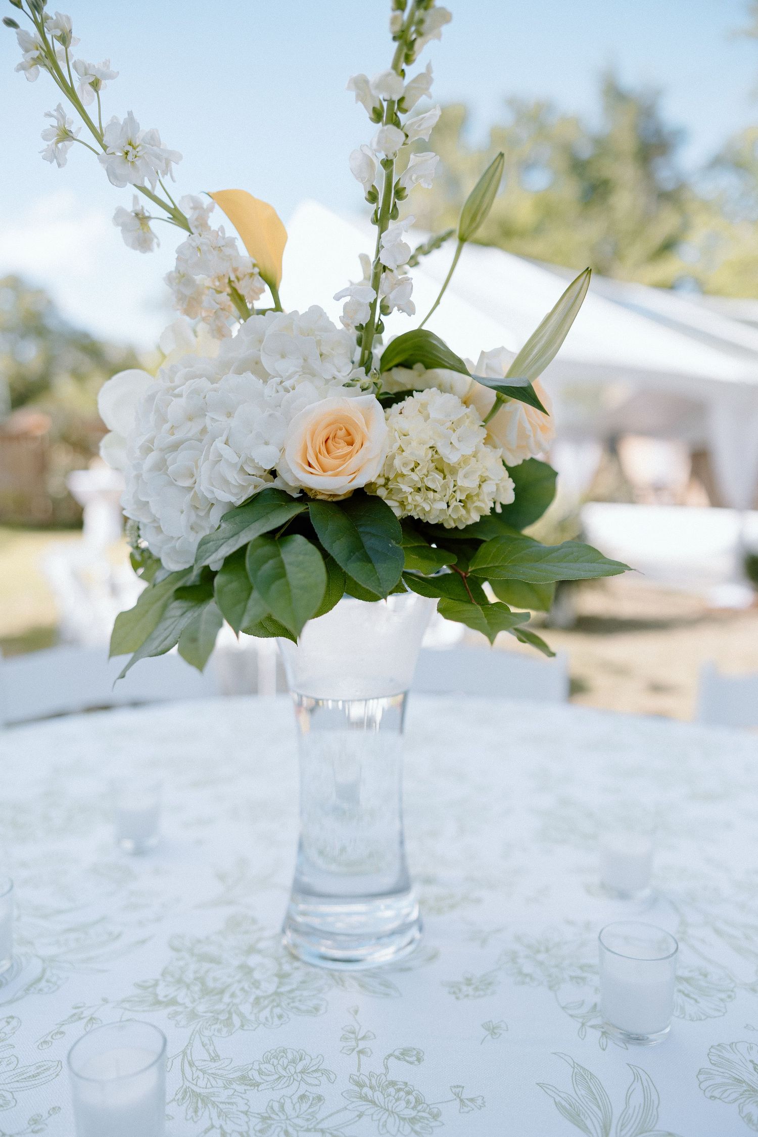 Elegant wedding centerpiece with white hydrangeas, peach roses, and greenery in glass vase.