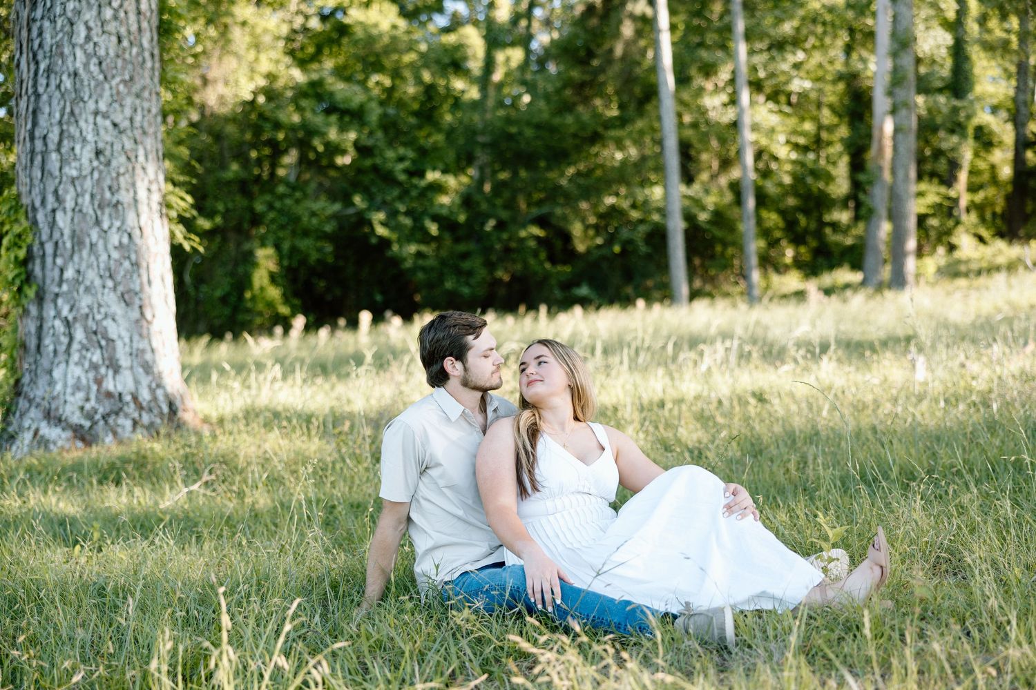 Couple sitting together in lush green grass field, leaning close and gazing at each other near tall trees.