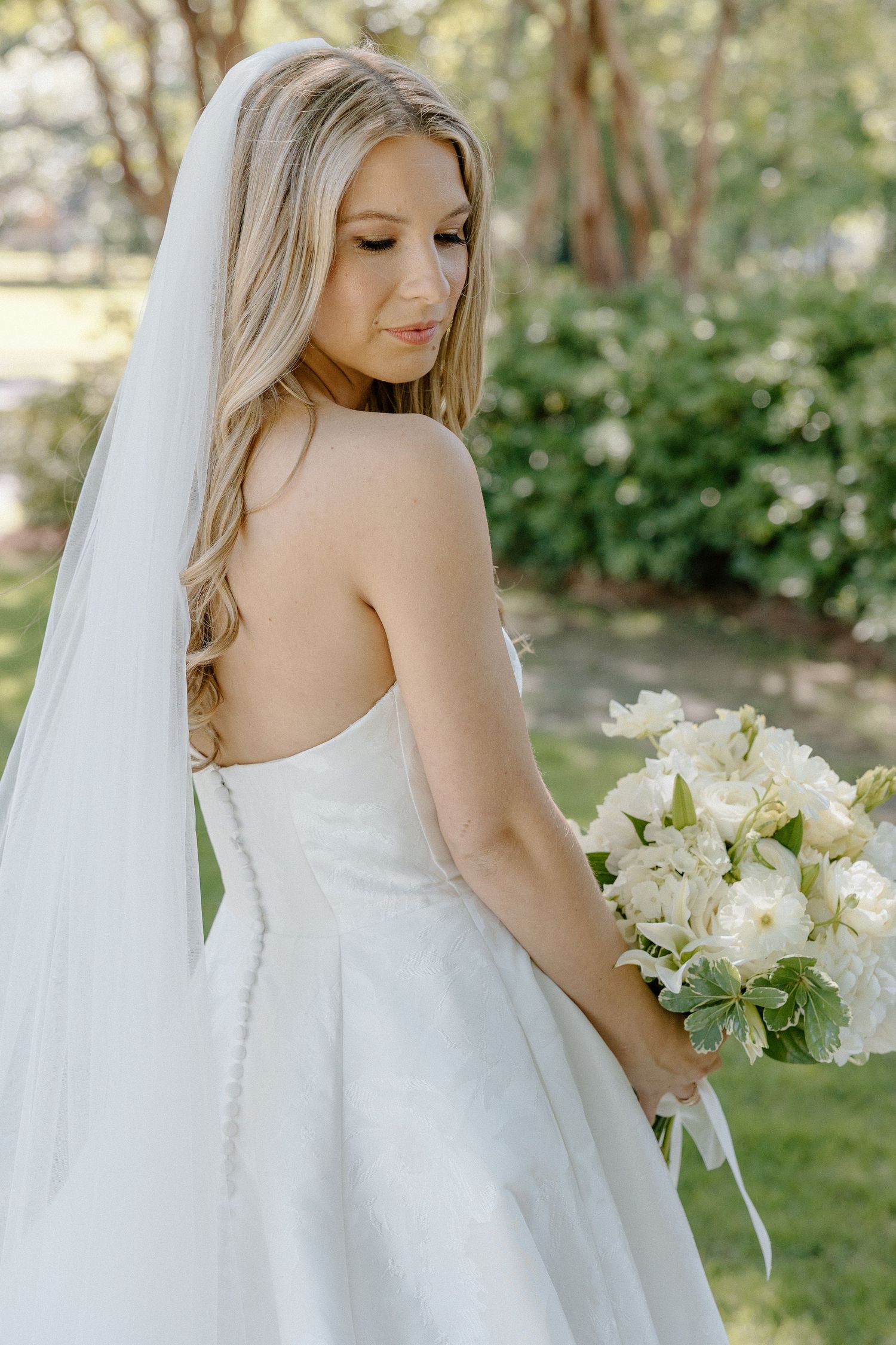 Bride in strapless white gown and veil holding white bouquet in lush garden setting.