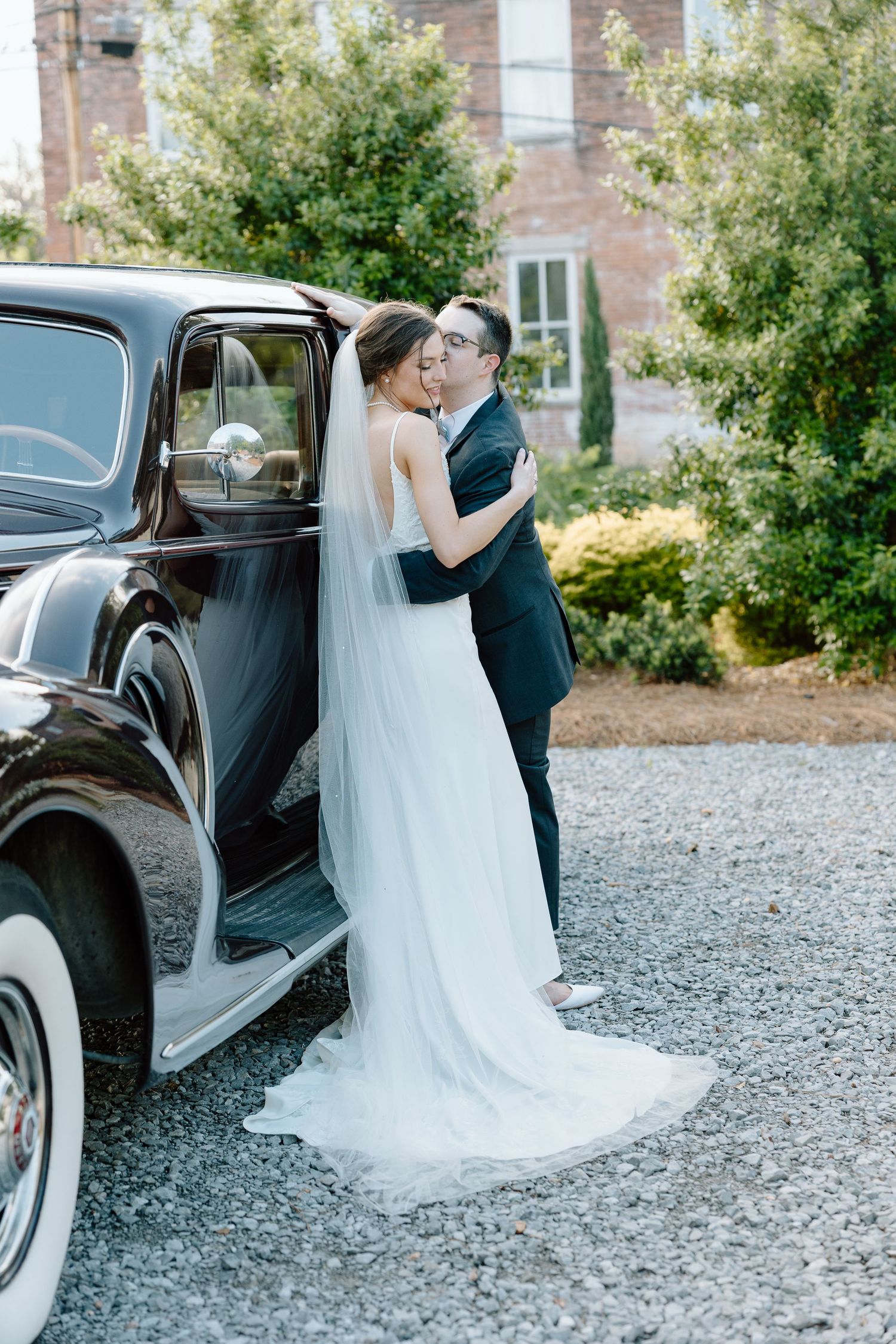 Bride and groom share a kiss beside a vintage black classic car in a lush garden setting.