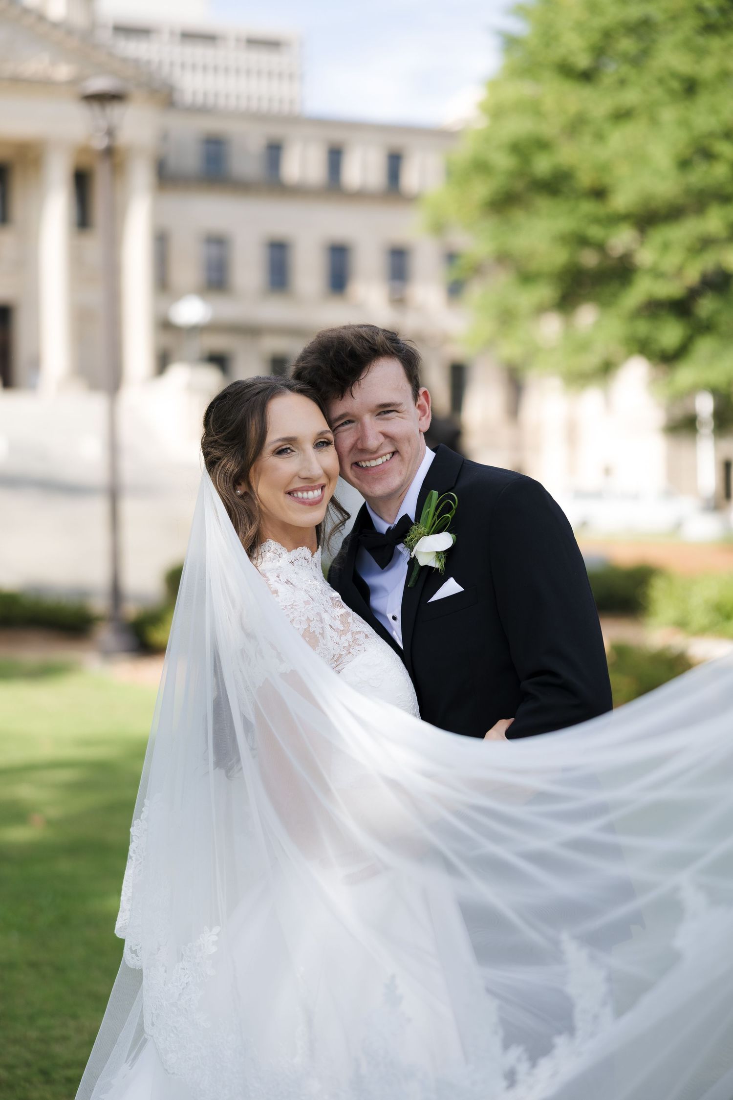 Smiling bride and groom pose outdoors, her lace wedding dress veil flowing in the wind, with a classic building backdrop.