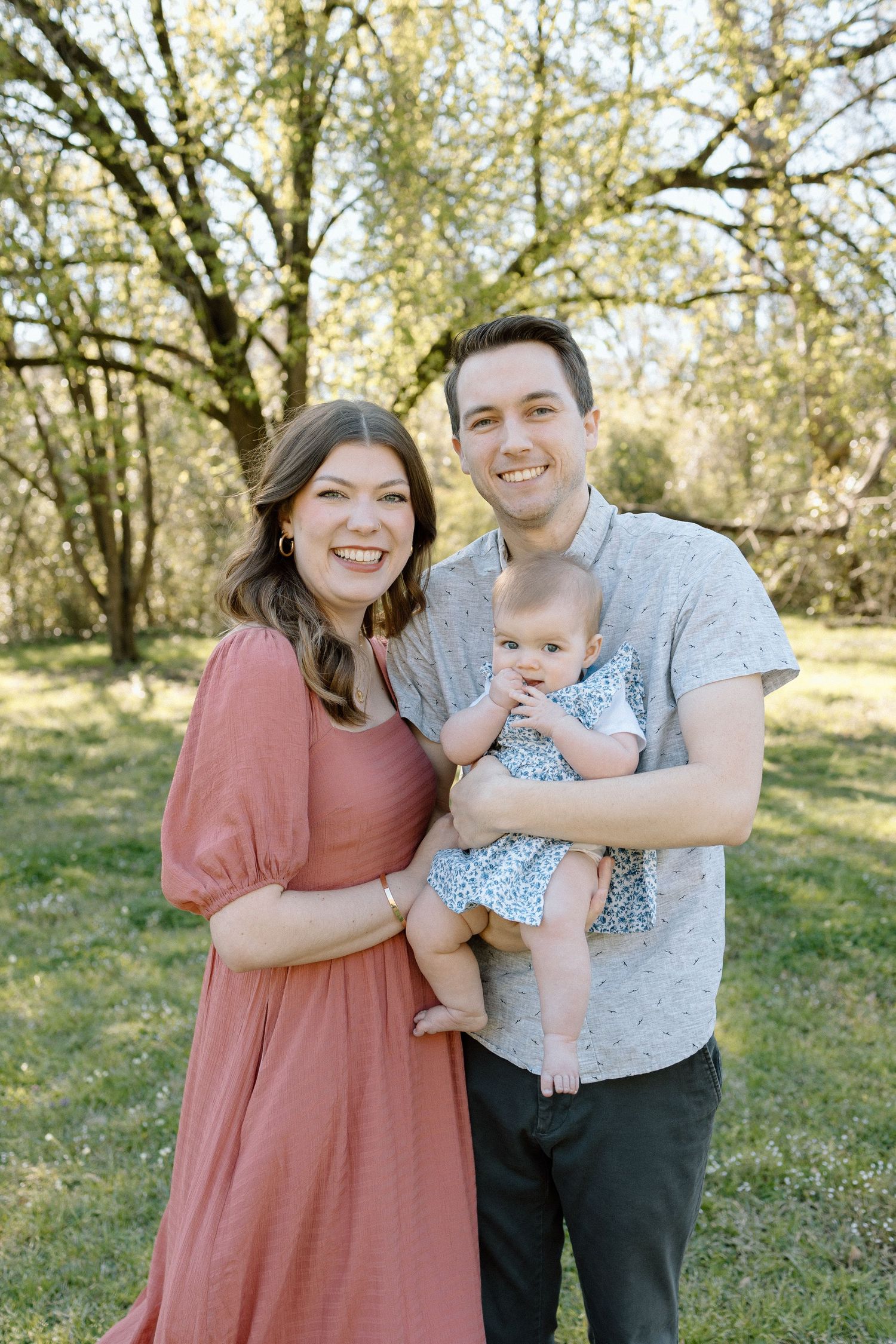 Smiling young family with mother in rust dress and father holding baby girl in spring outdoor portrait.