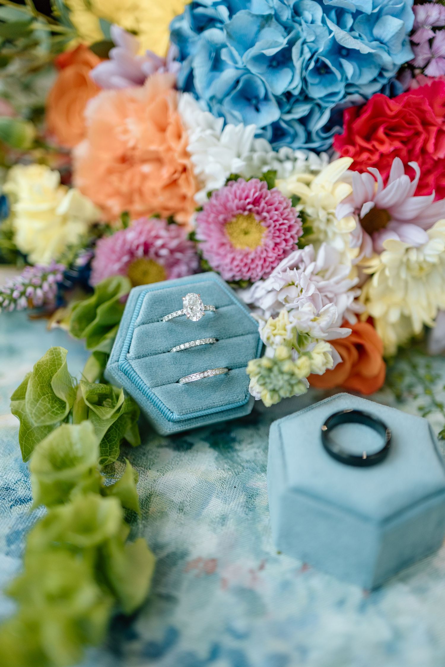 Elegant wedding rings displayed in a blue velvet hexagon box surrounded by colorful spring flowers.