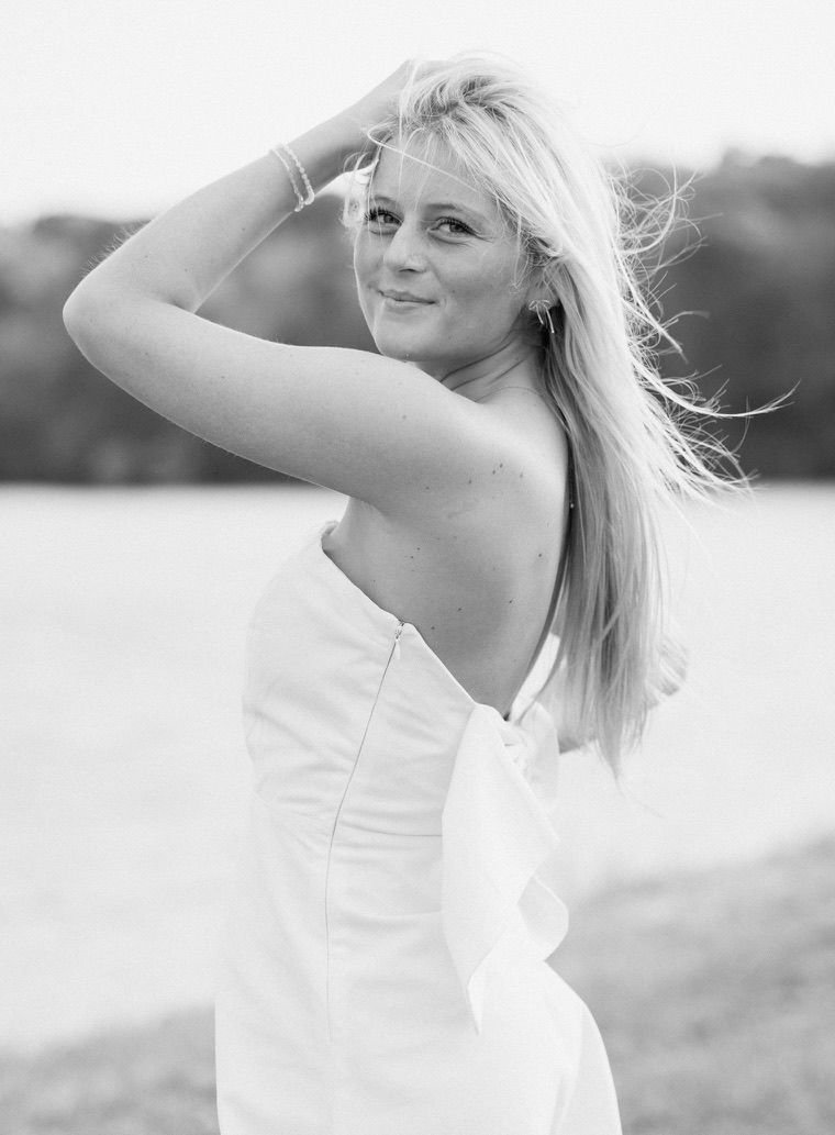 Blonde woman in white backless dress posing near waterfront in black and white photo.