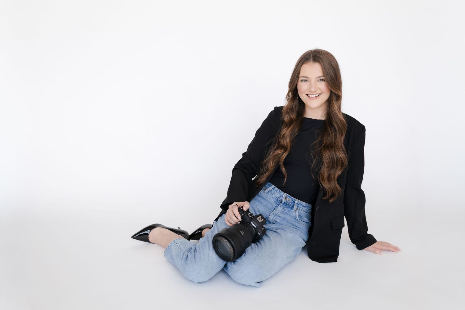 Young woman with long brown hair sits on white floor holding a DSLR camera, wearing black blazer and jeans.