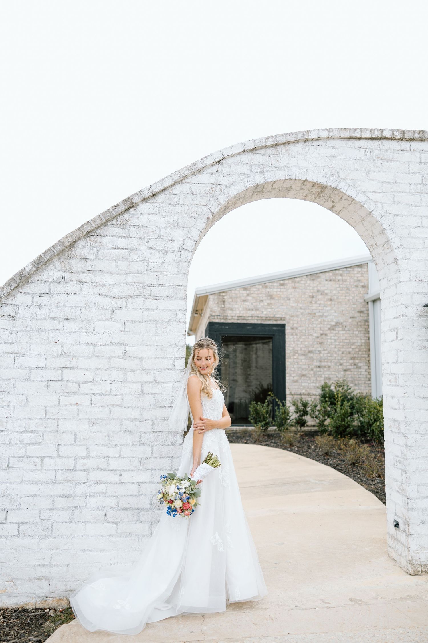 Smiling bride in white gown holding bouquet under white brick archway at venue.