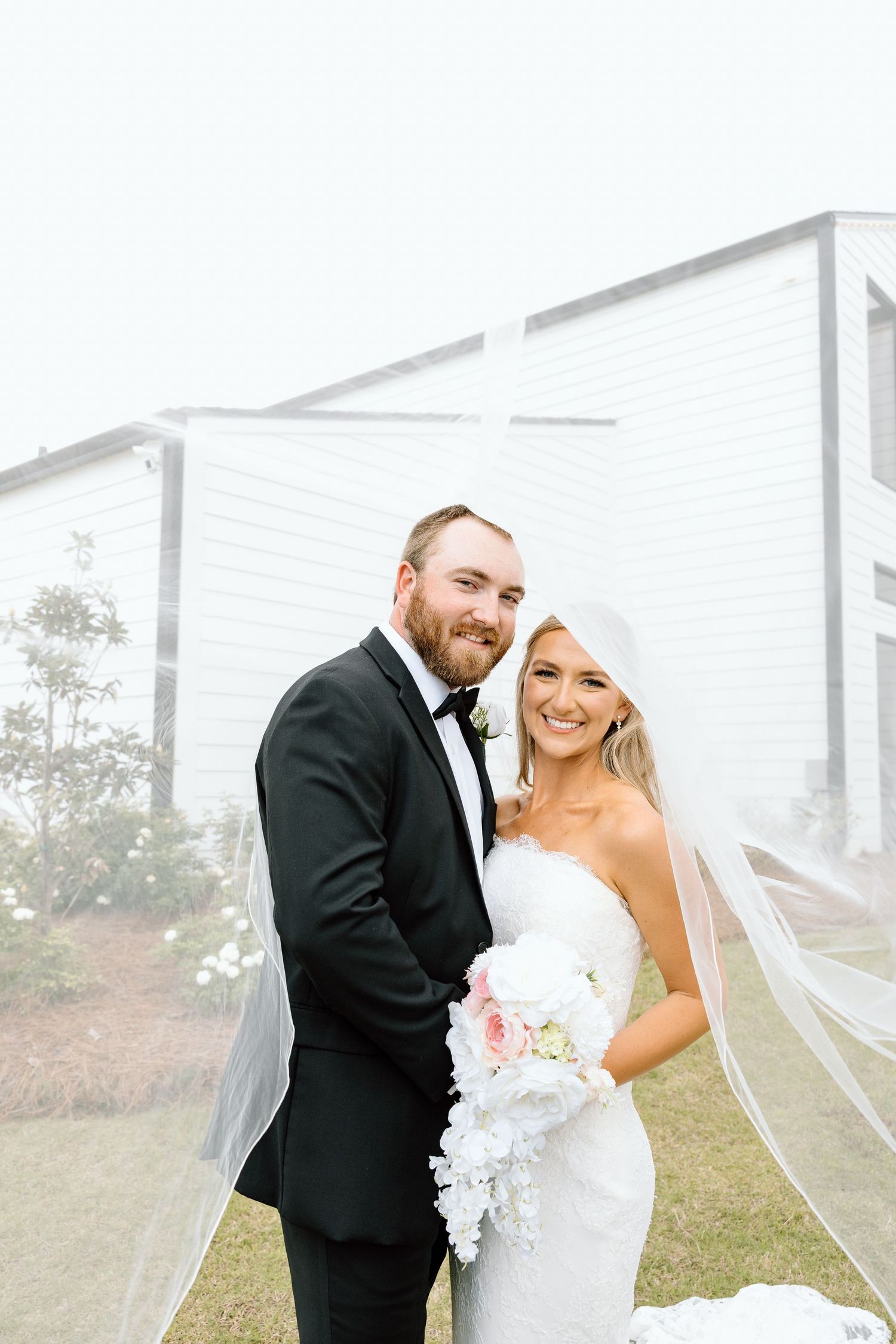 Smiling bride and groom pose together in front of white cross at outdoor wedding venue on misty day.