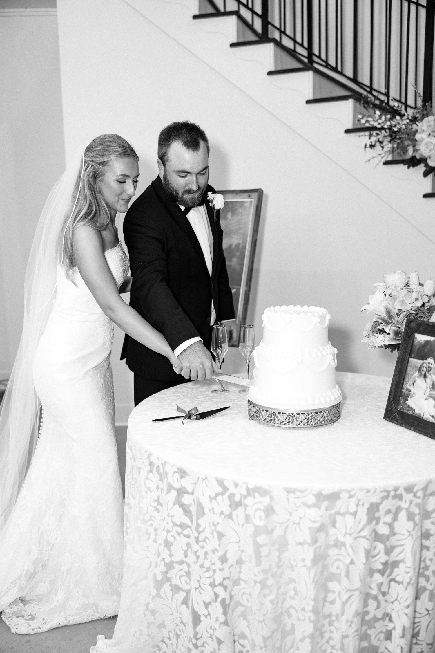 Bride and groom cut elegant two-tier white wedding cake together in black and white photo indoors.