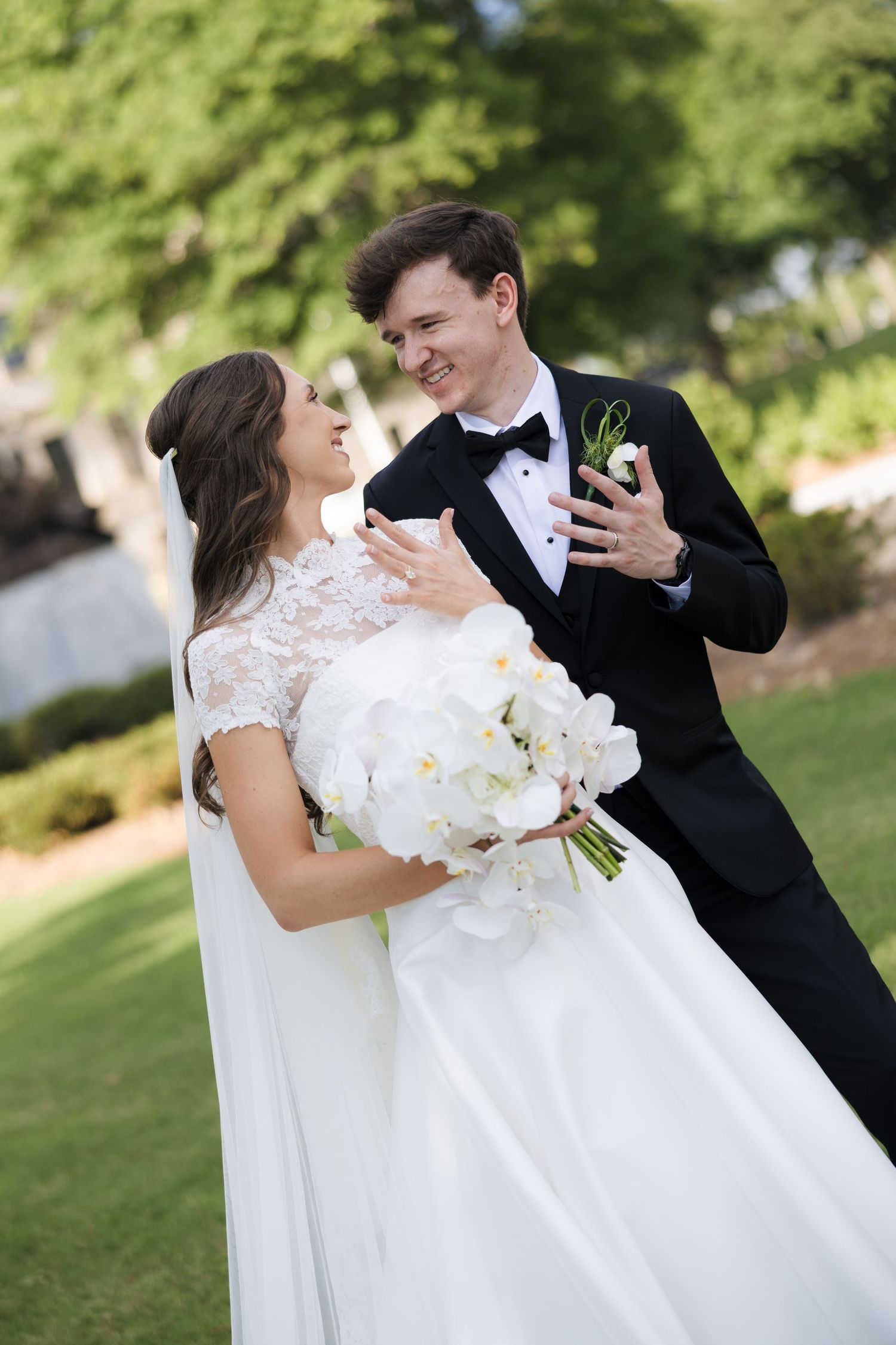 Smiling bride and groom in wedding attire share a joyful moment outdoors, bride holding white orchid bouquet.
