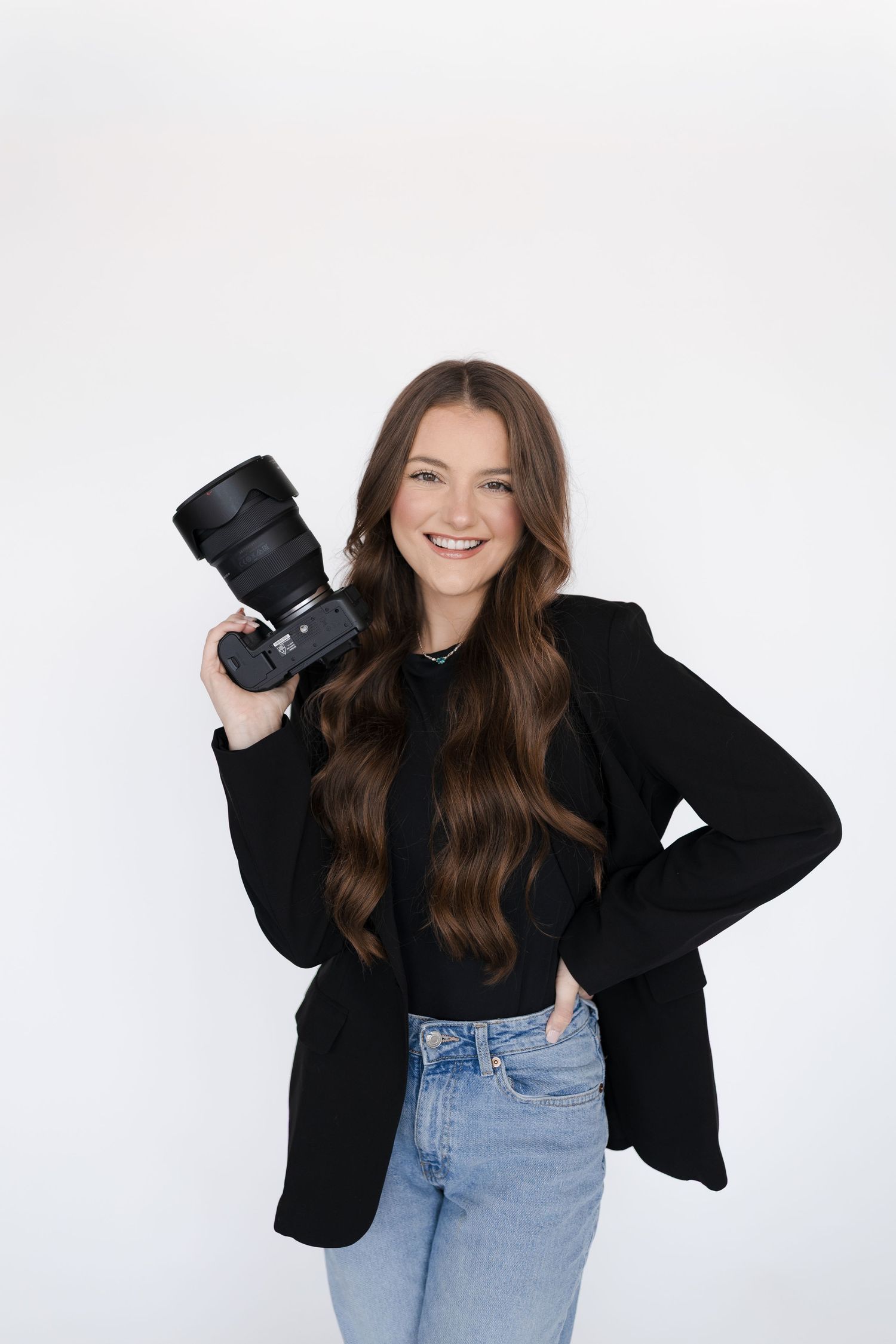 Smiling young woman with long brown hair holding a DSLR camera, wearing black blazer and jeans on white background.