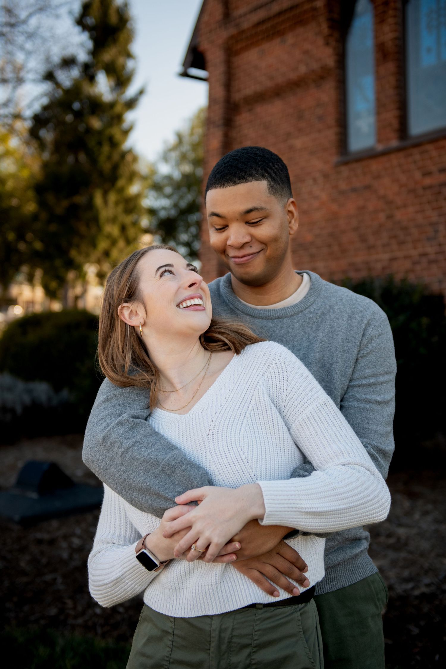 A couple embraces outdoors in front of a brick building, sharing a tender moment in warm sunlight.