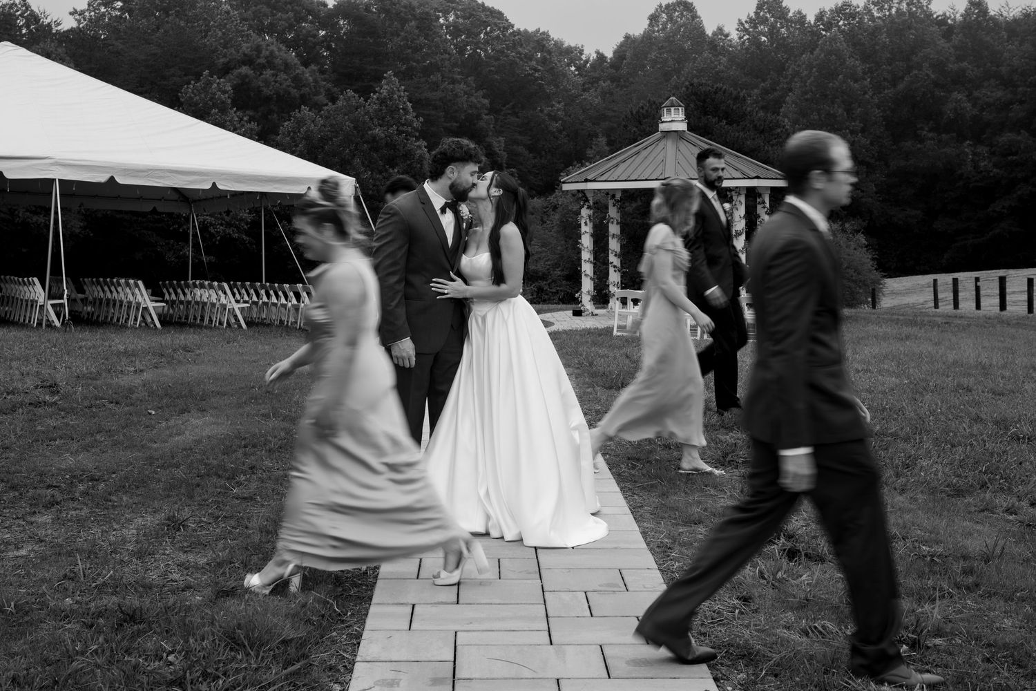 Black and white photo of bride and groom kissing surrounded by wedding party on brick path at outdoor venue.