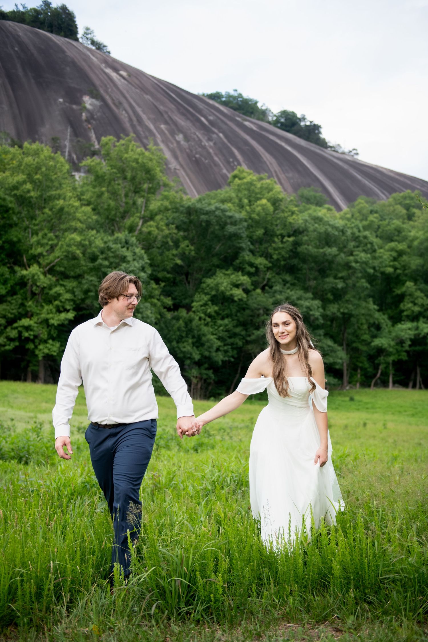 Couple holding hands walking through green meadow with large granite rock formation and trees in background.