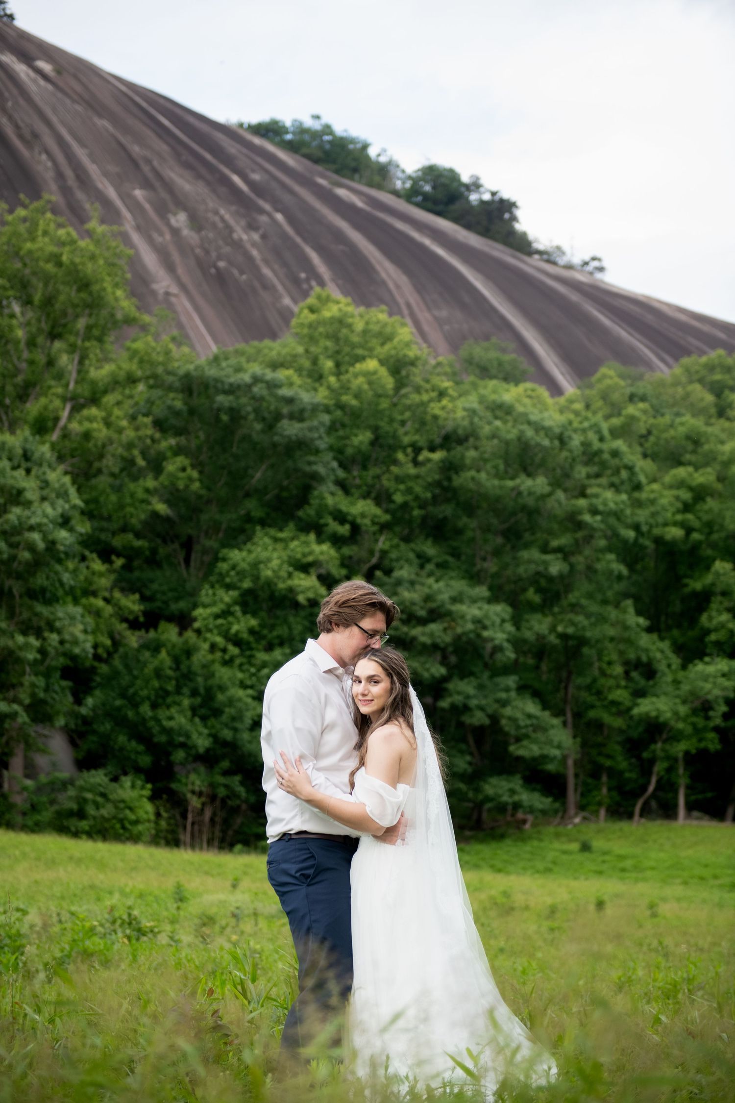 Bride and groom embracing in a green meadow with lush trees and a large granite rock formation in the background.