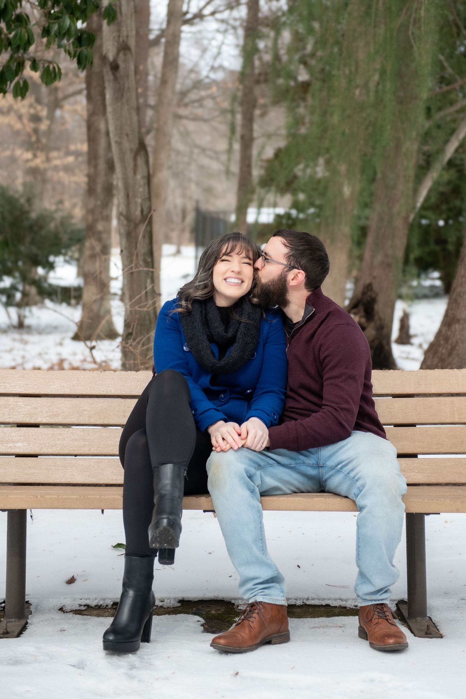 A couple shares a tender moment while sitting together on a wooden bench in a snowy park.