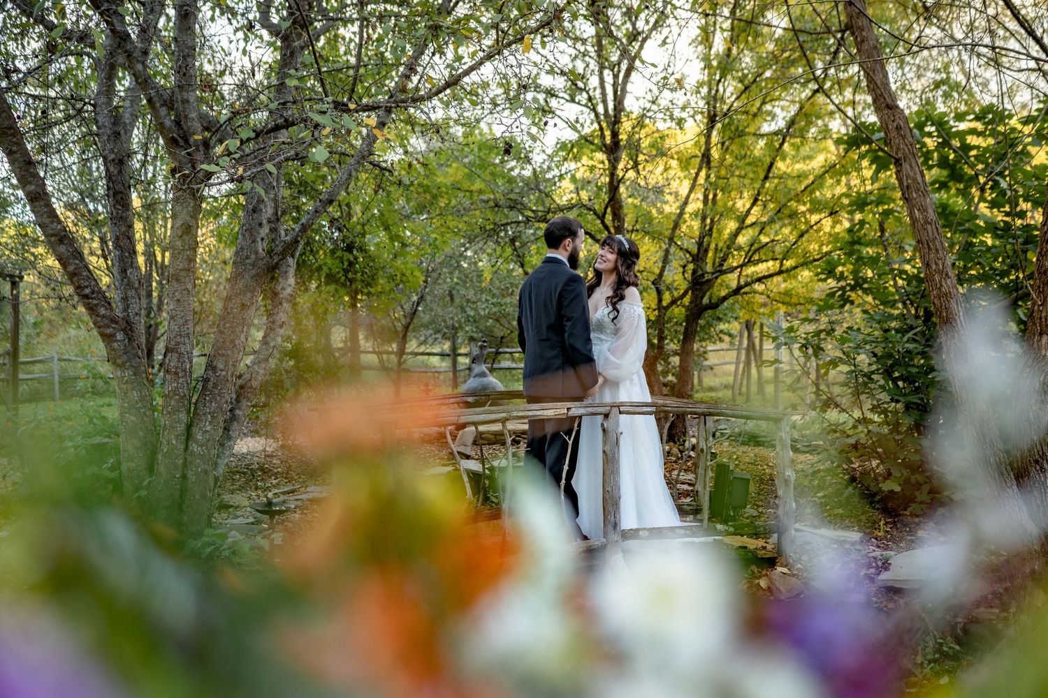Bride in a white gown and groom in a dark suit embrace on a wooden bridge surrounded by trees, framed by blurred colorful flowers.