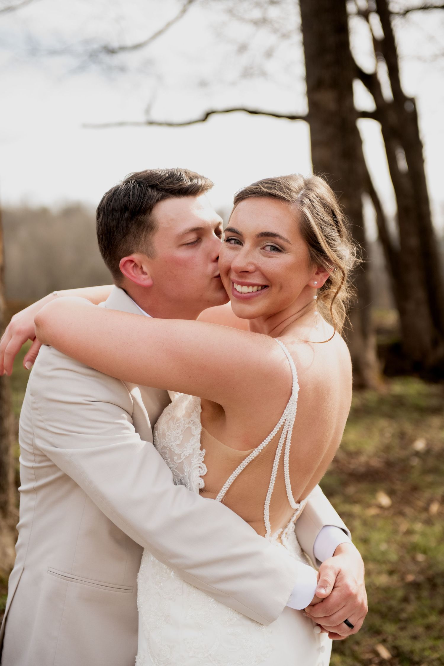 A groom in a tan suit kisses his smiling bride on the cheek outdoors, as she wears a backless lace wedding gown.