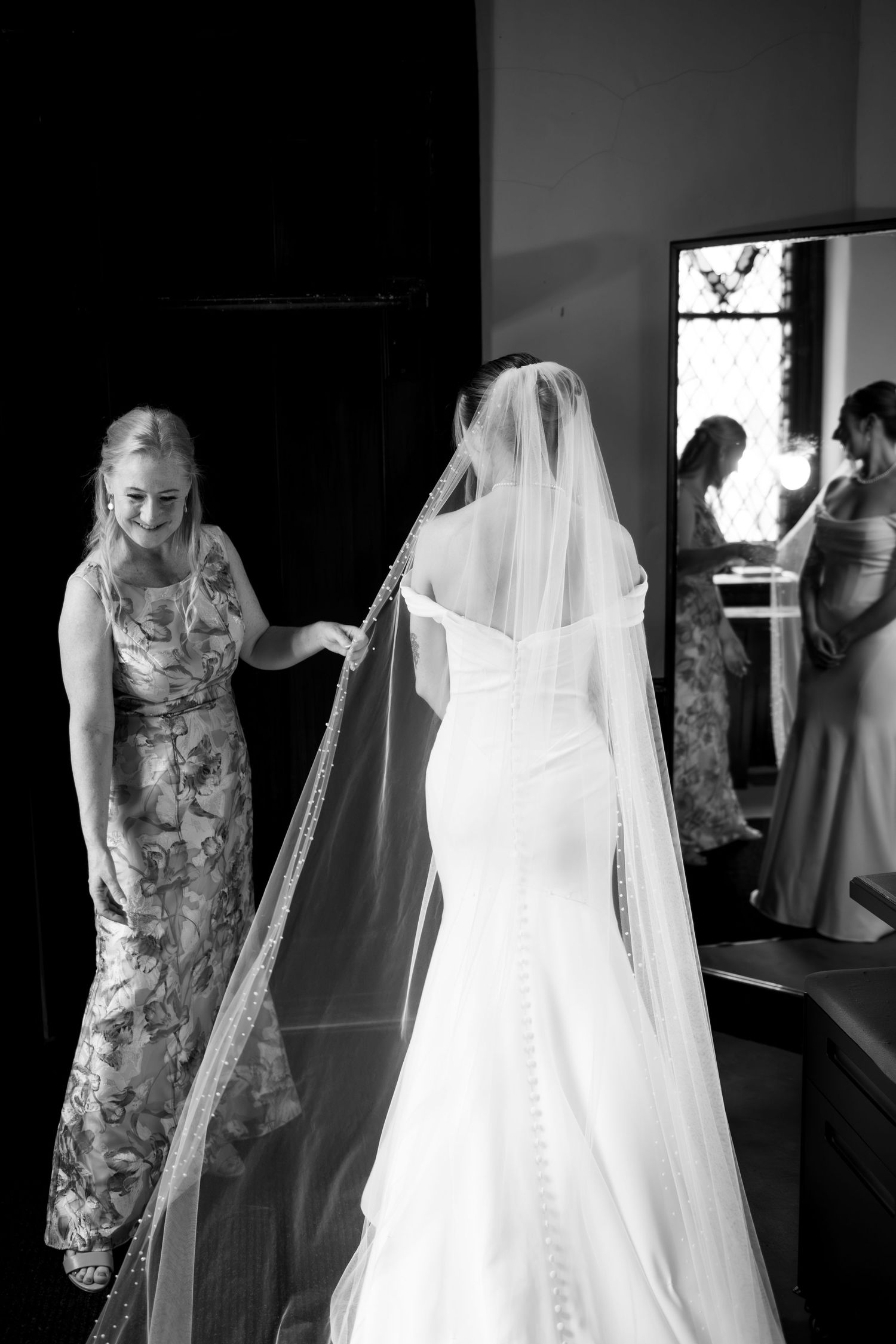 Black and white photo of a bride in a fitted gown and veil being assisted by a smiling bridesmaid before the wedding ceremony.