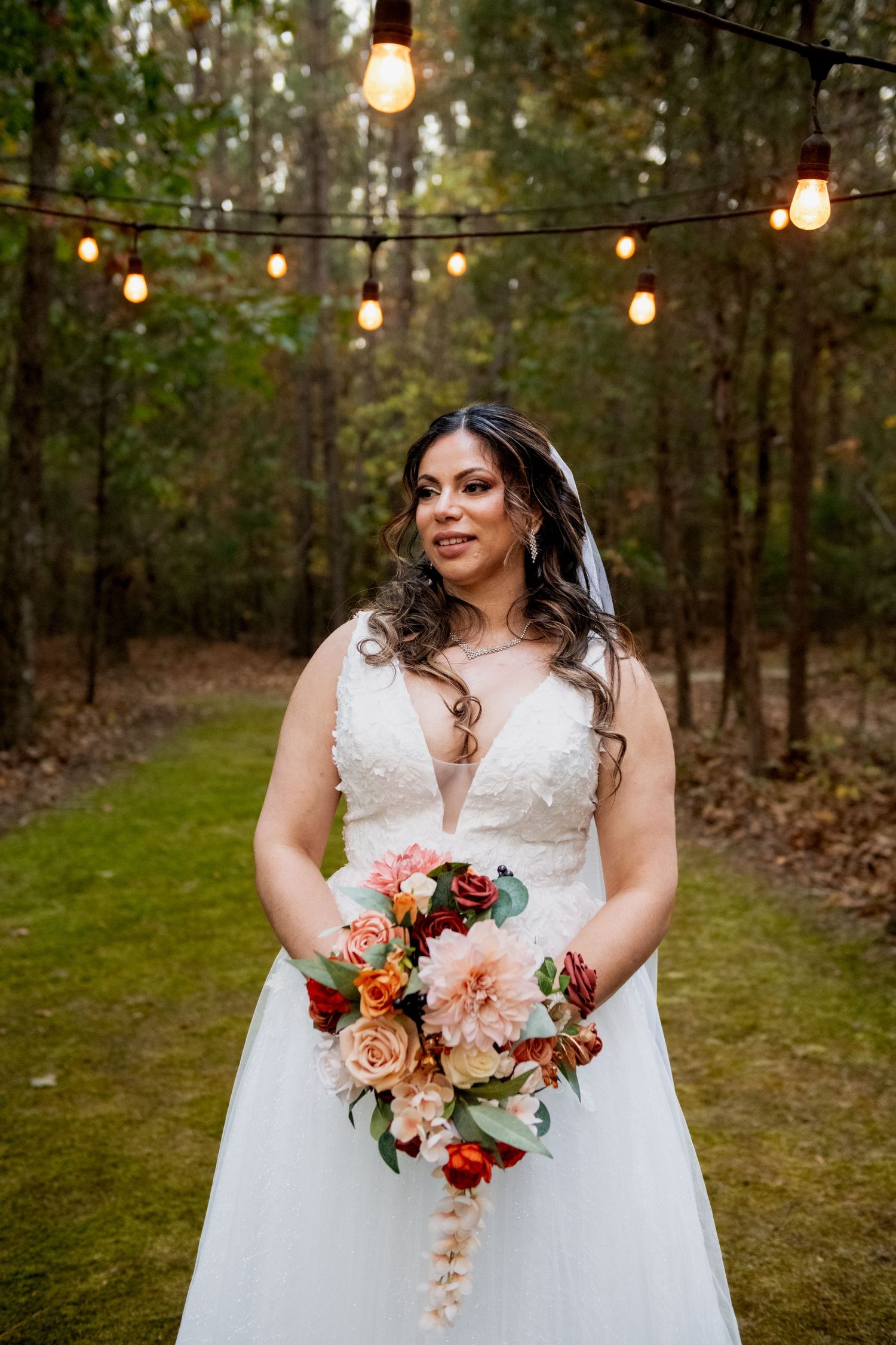 Beautiful bridal bouquet with coral and peach flowers held against a white wedding dress in an outdoor woodland setting.